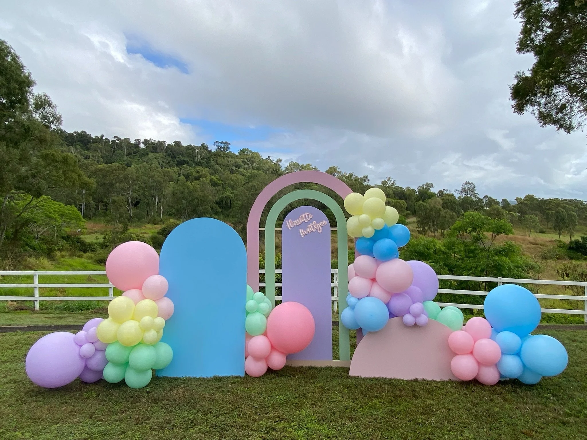 Colorful balloon arrangement with pastel pink, yellow, green, purple, and blue balloons in front of pastel purple, blue, and pink arches on grass with trees and cloudy sky in the background.