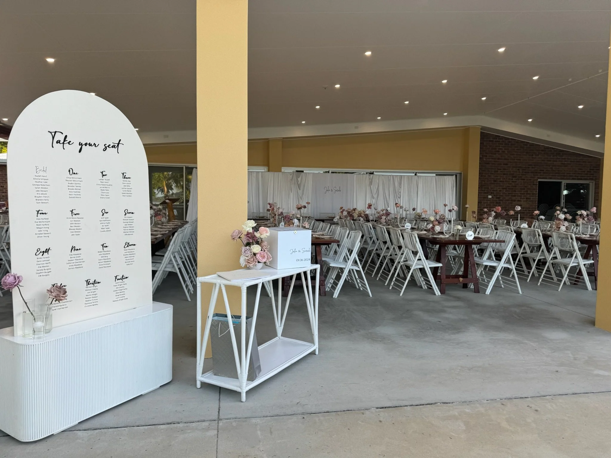 Wedding reception setup with decorated tables, chairs, and floral arrangements, with a large seating chart display and a sign with the couple's names.