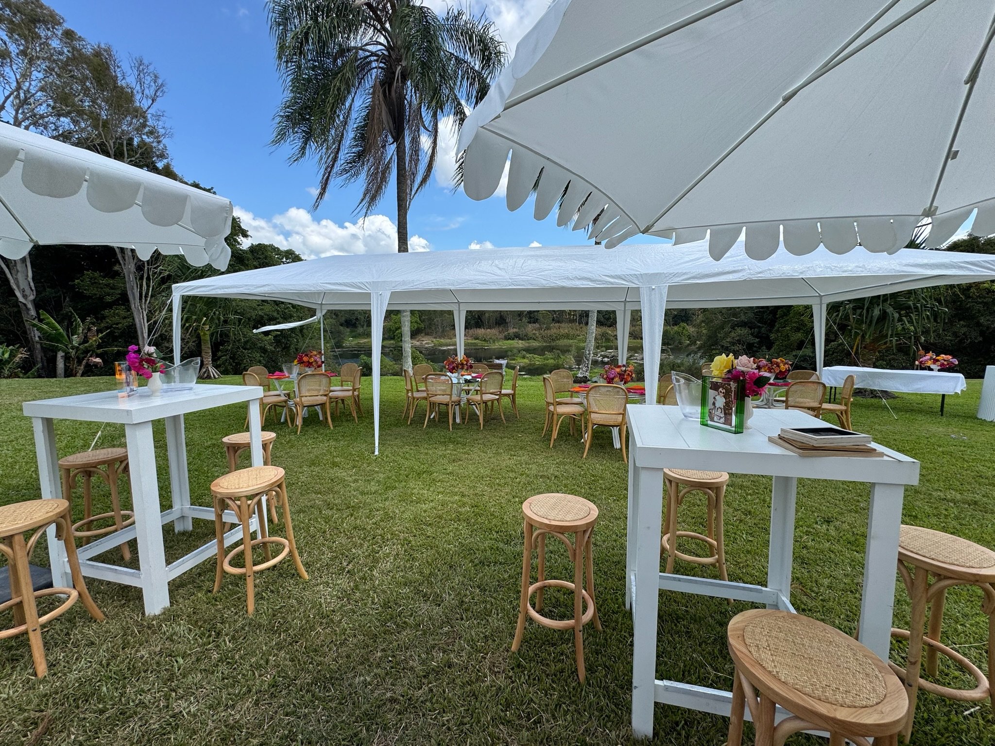 Outdoor event setup with white tents, round wooden tables with floral centerpieces, bar-height white tables with chairs, and white umbrellas, on a grassy lawn near a small lake with trees and a partly cloudy sky.