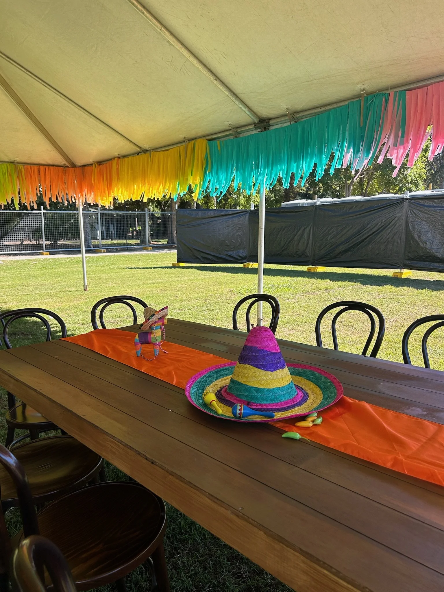 Decorated outdoor party table with colorful sombrero hats and balloons on an orange table runner, under a tent with rainbow fringe decorations.