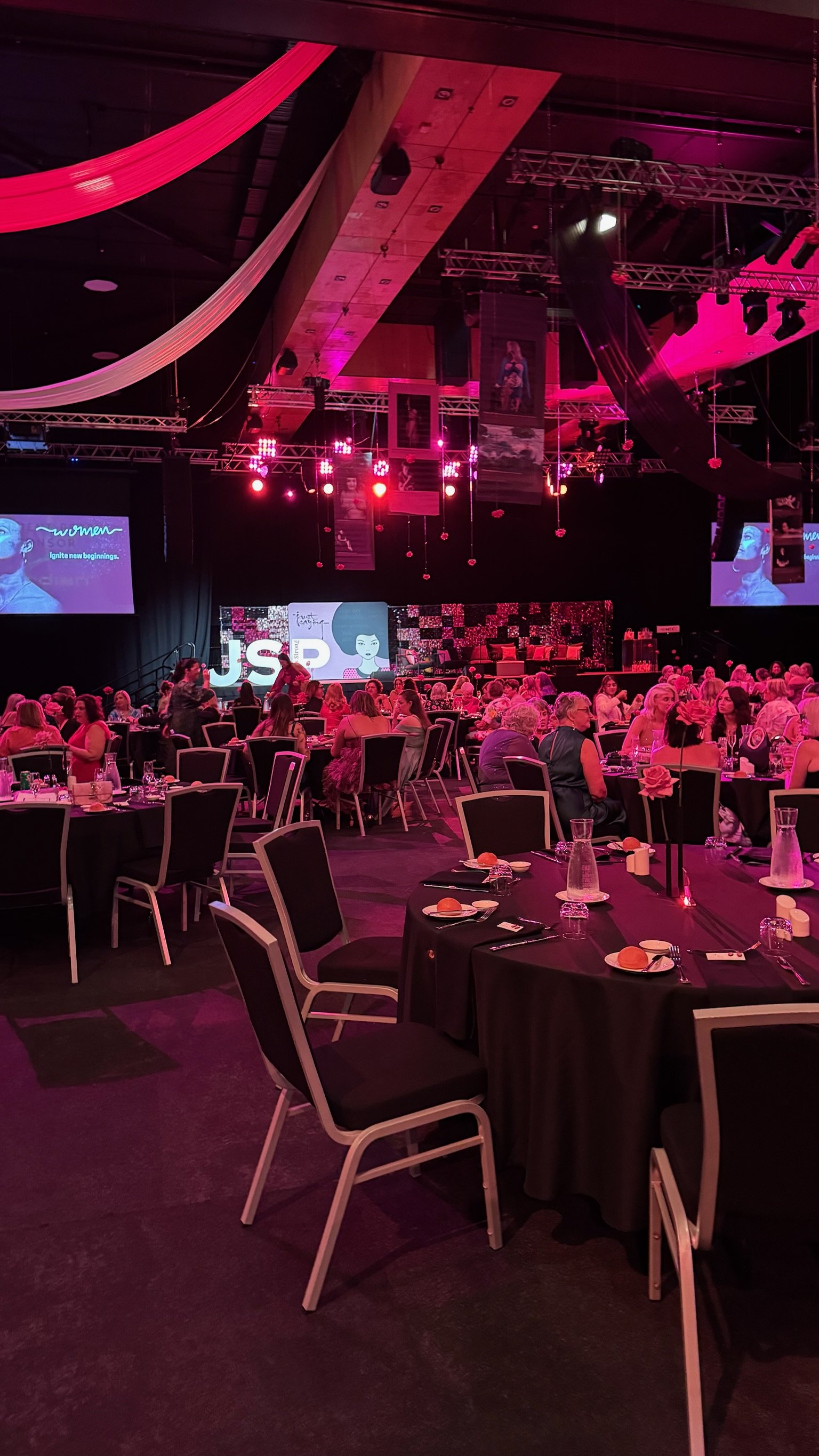 A large indoor event space decorated with pink and purple lighting, with round tables covered with black tablecloths, set with plates, glasses, and forks. There are many people seated at the tables, facing a stage with a large screen displaying the w