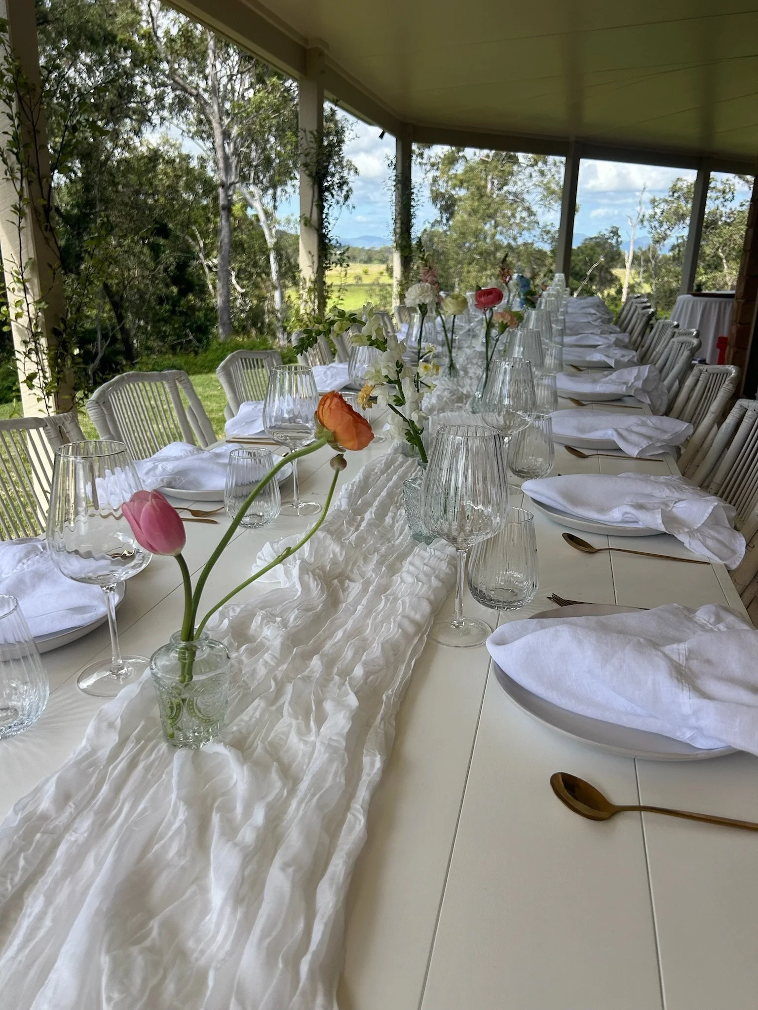 Long banquet table set for a formal event with white tablecloth, gold utensils, white napkins, and glassware, decorated with a white fabric runner and small vases of pink, orange, and white flowers. The setting is on a screened porch with trees and a