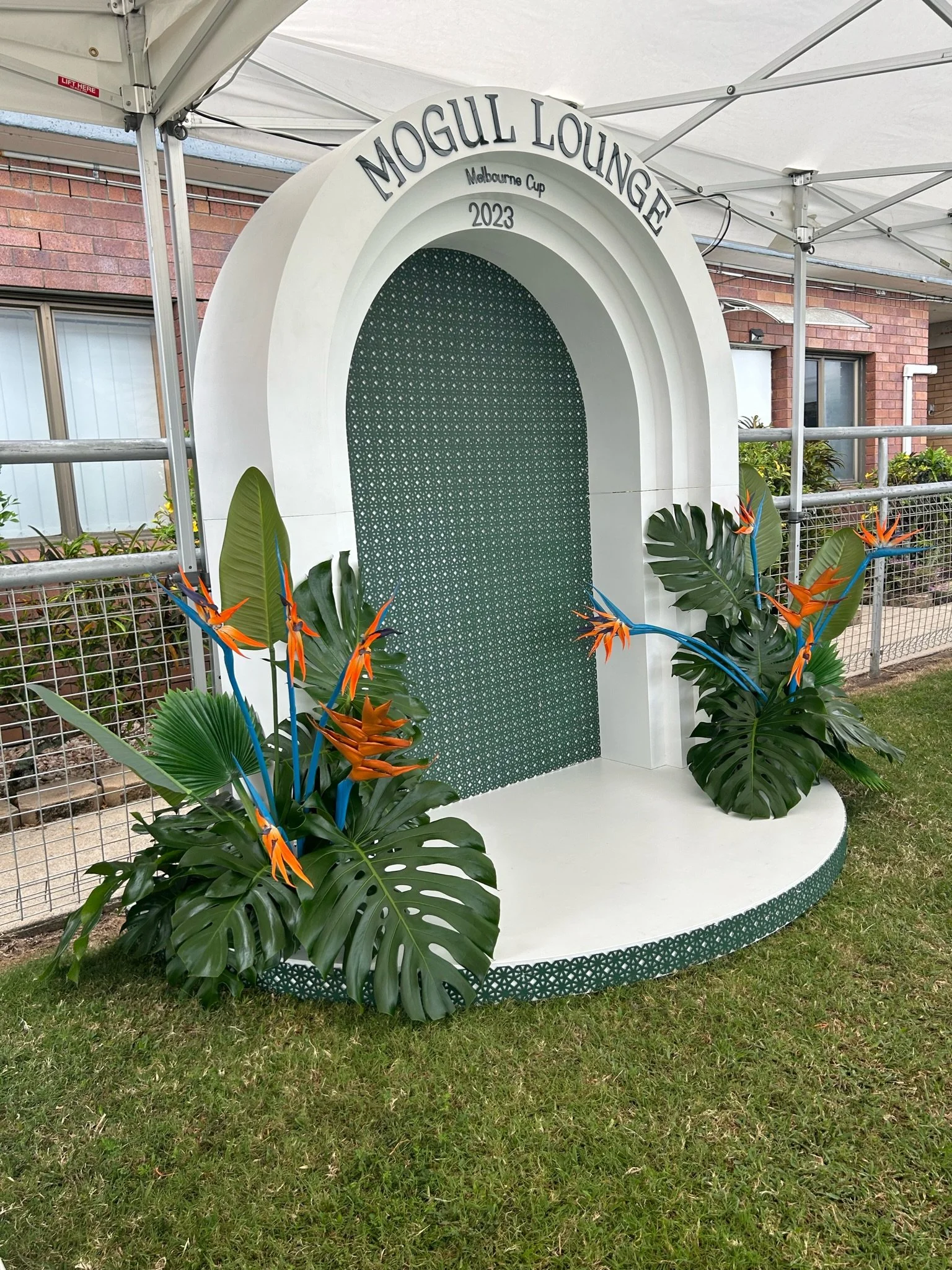Decorative arch with green foliage, orange and blue flowers, and signage reading "Mogul Lounge Melbourne Cup 2023".