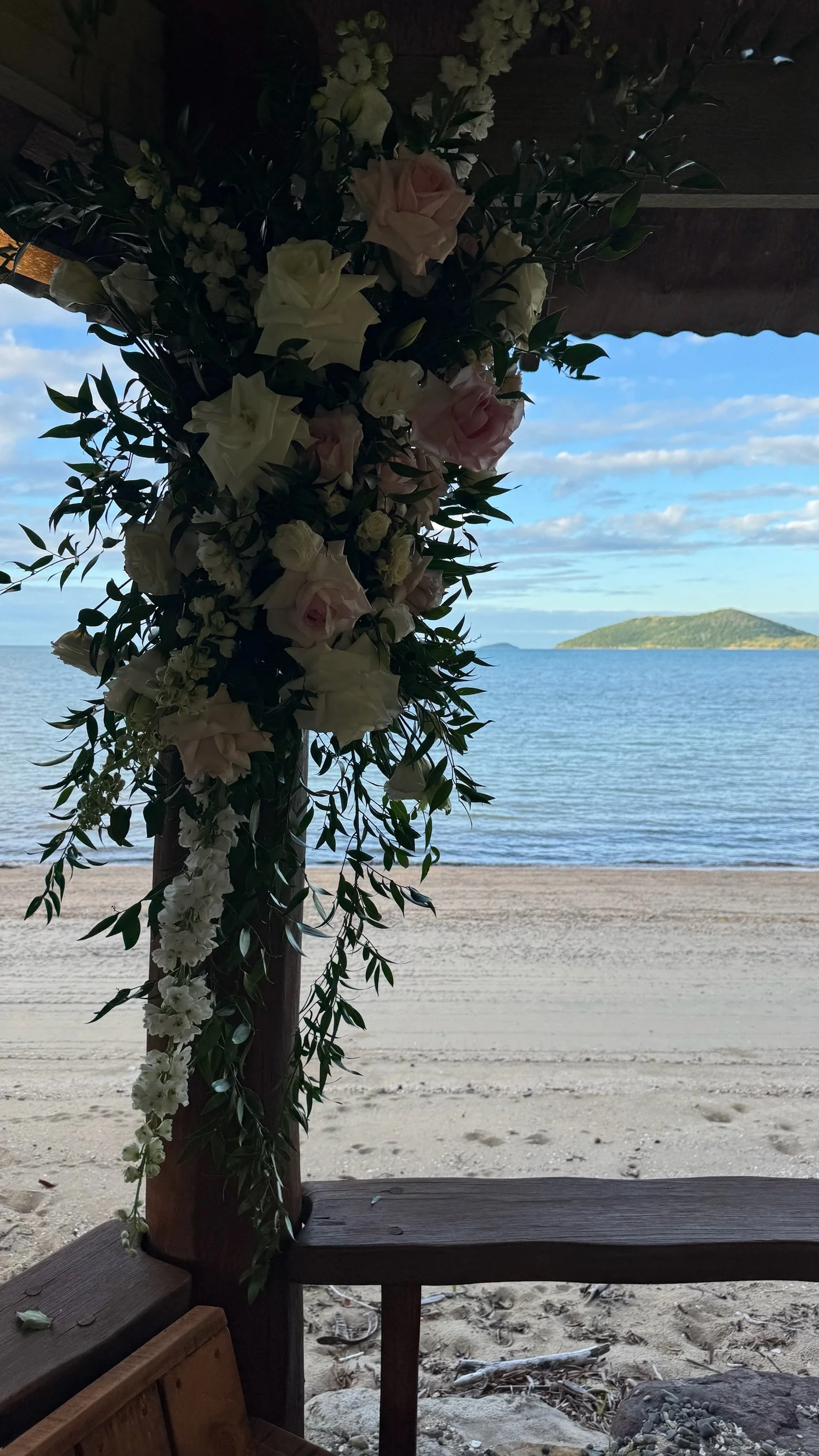 Flower arrangement with white and pink roses and greenery hanging from a wooden structure at a beach, with sandy shore, water, and island in the background.