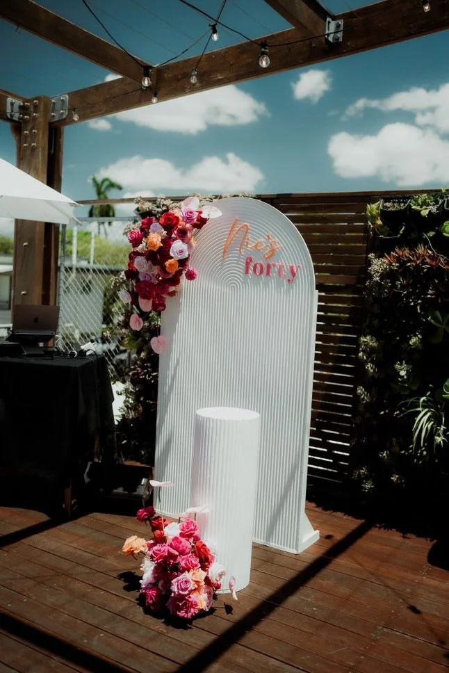 A floral backdrop with pink and red flowers, a white curved backdrop with the words "Not foray" written in orange and pink, a white pedestal, and a bouquet of pink flowers on a wooden patio under a blue sky with clouds.