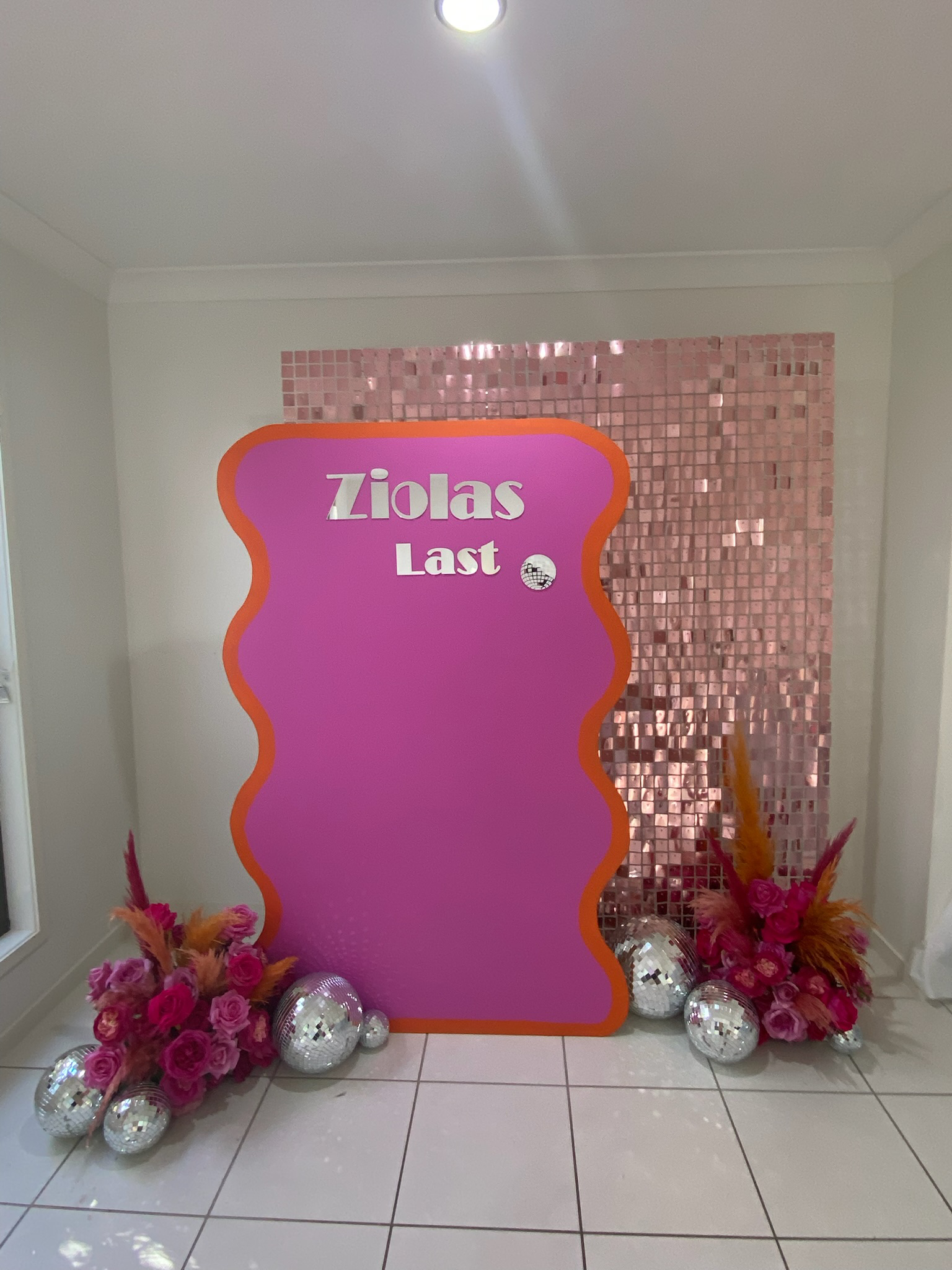 Decorative pink backdrop with the words 'Ziolas Last' in large white letters, surrounded by pink flowers and silver disco balls, set against a pink mosaic wall.