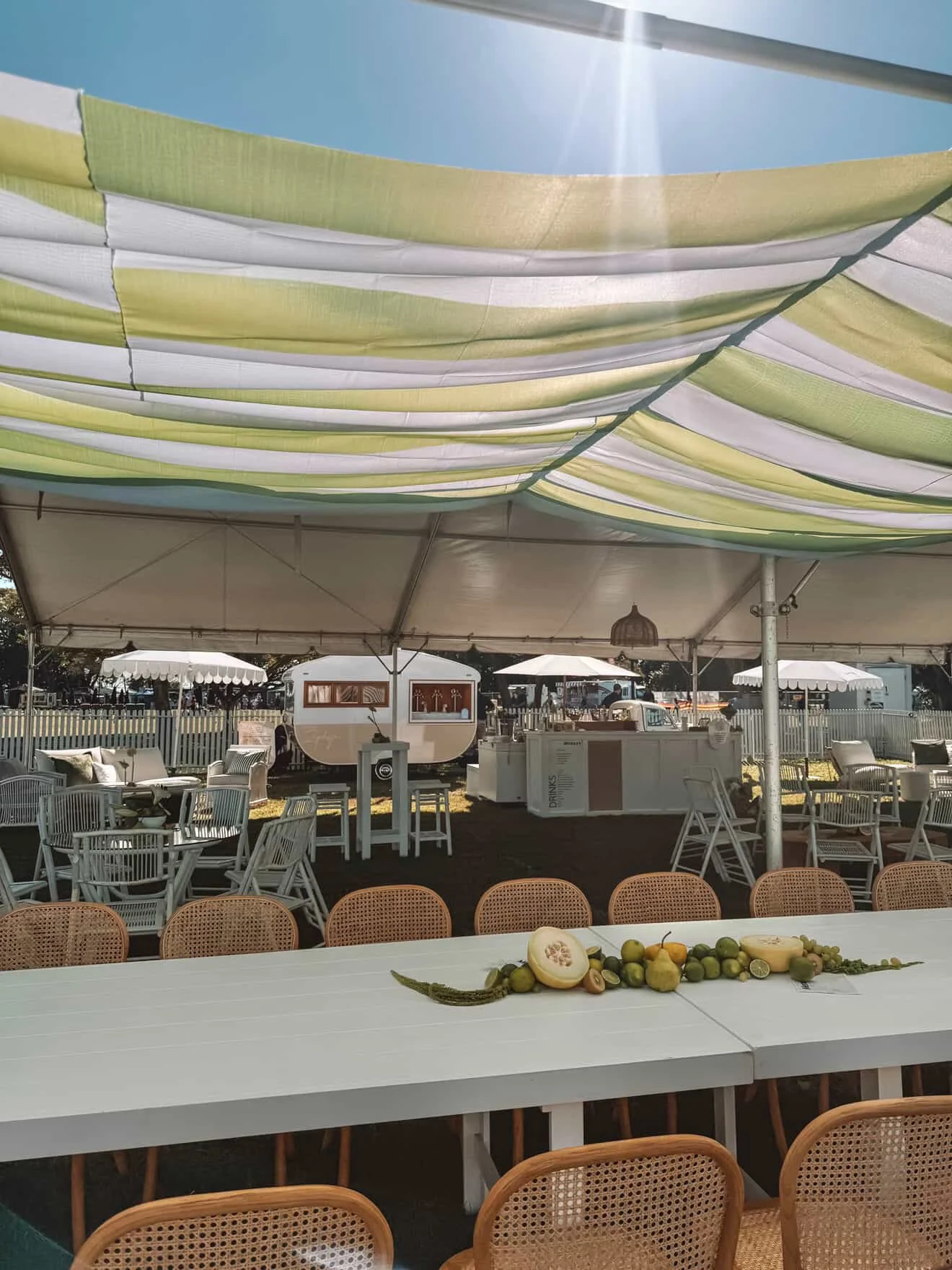 Outdoor event setup under a large, striped canopy with tables, chairs, and a decorative arrangement of fruit and vegetables on a white table in the foreground.