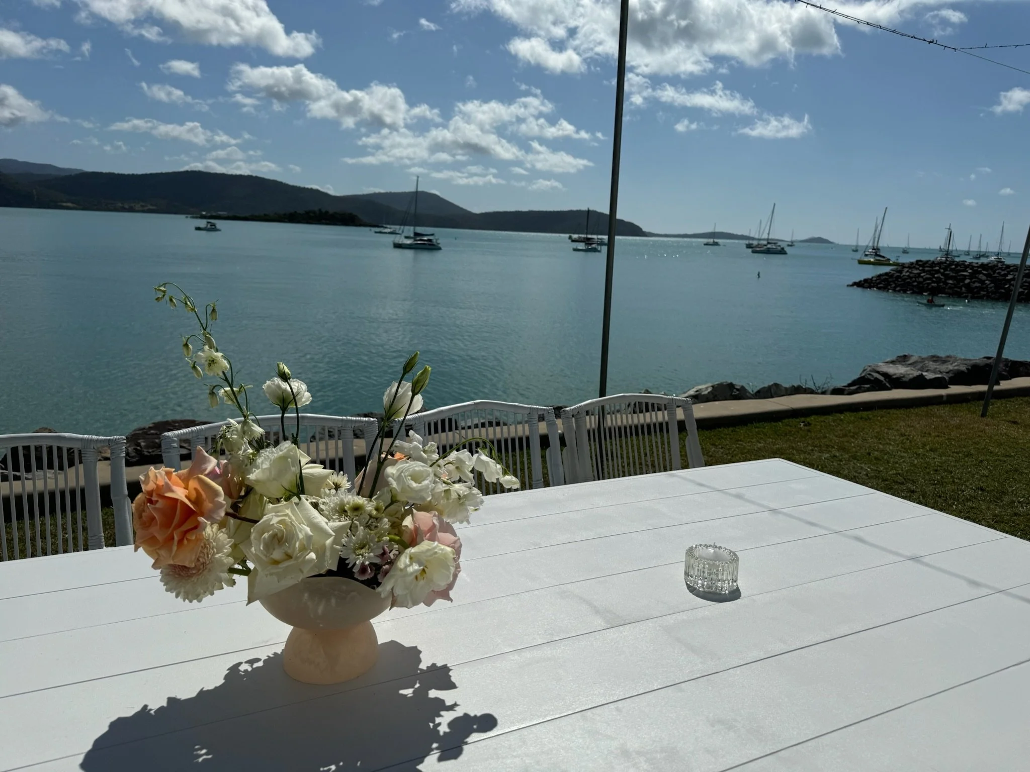 A white outdoor table with a vase of white and peach flowers, and a small glass candle holder, overlooking a bay with sailboats and a mountainous shoreline under a partly cloudy sky.