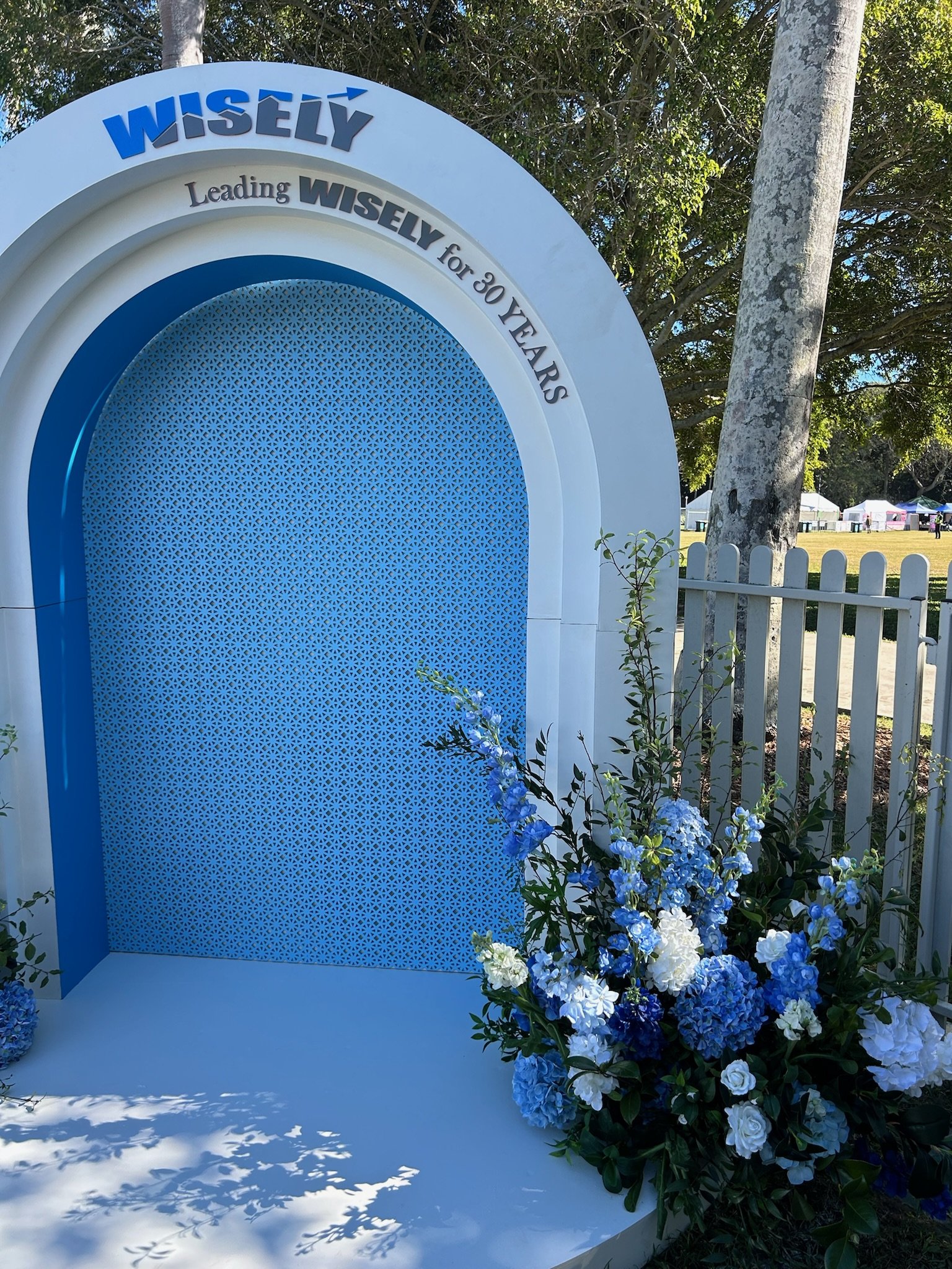 Blue and white display with floral arrangement celebrating Wisely's 30-year milestone, featuring an arched design with the words 'Leading Wisely for 30 Years' and decorative background.