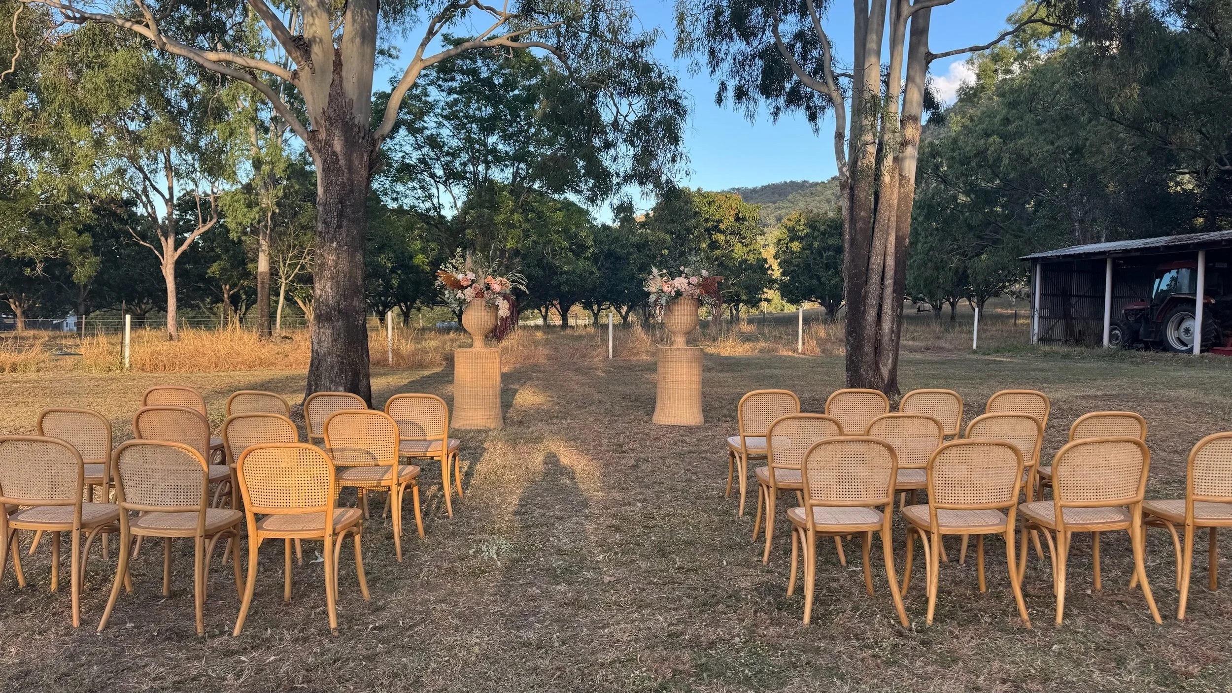 Outdoor setup with multiple beige chairs arranged in rows on grass, two large floral arrangements in vases on tall wicker stands, trees, a fence, and a shed with tractor in the background, under a clear sky.