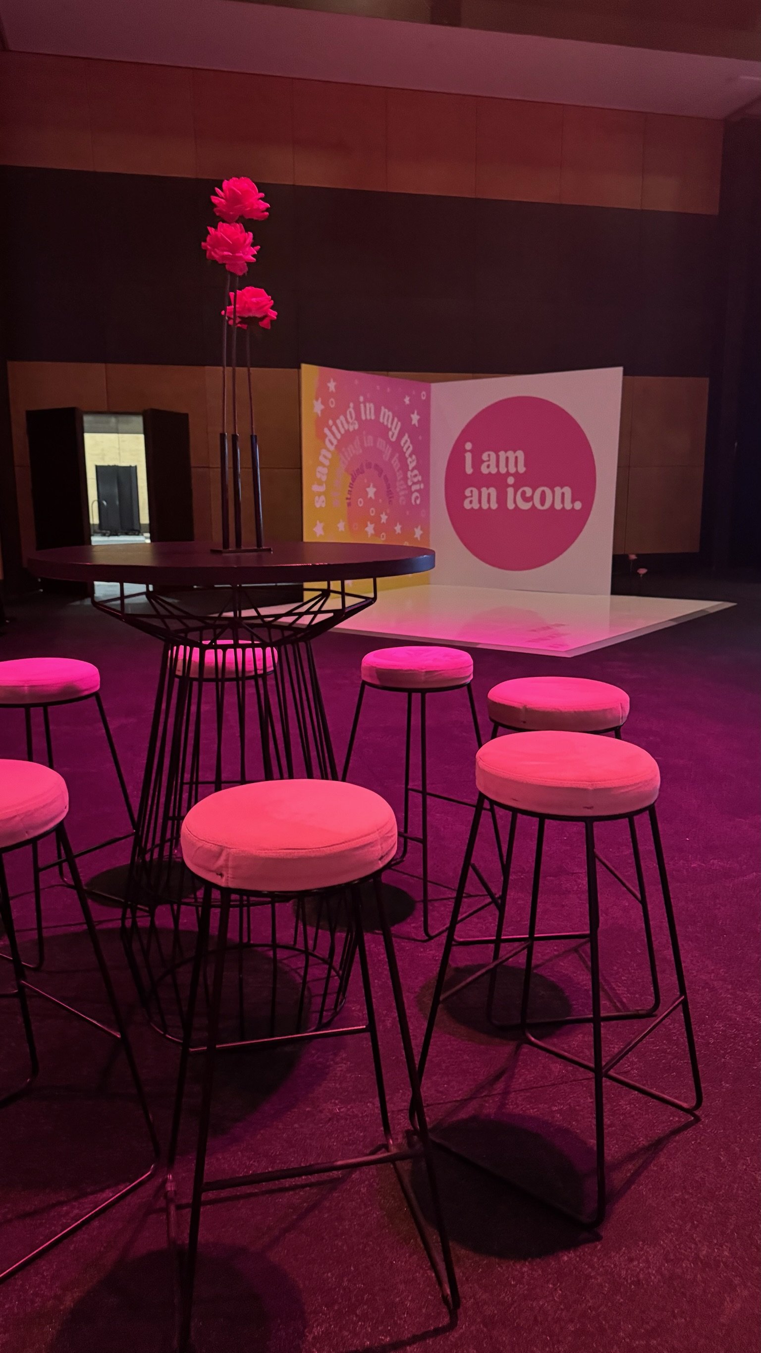 Round table with pink flower centerpiece surrounded by six pink-cushioned bar stools, set in a dimly lit indoor space with a backdrop featuring colorful signs that say 'standing in my magic' and 'I am an icon'.