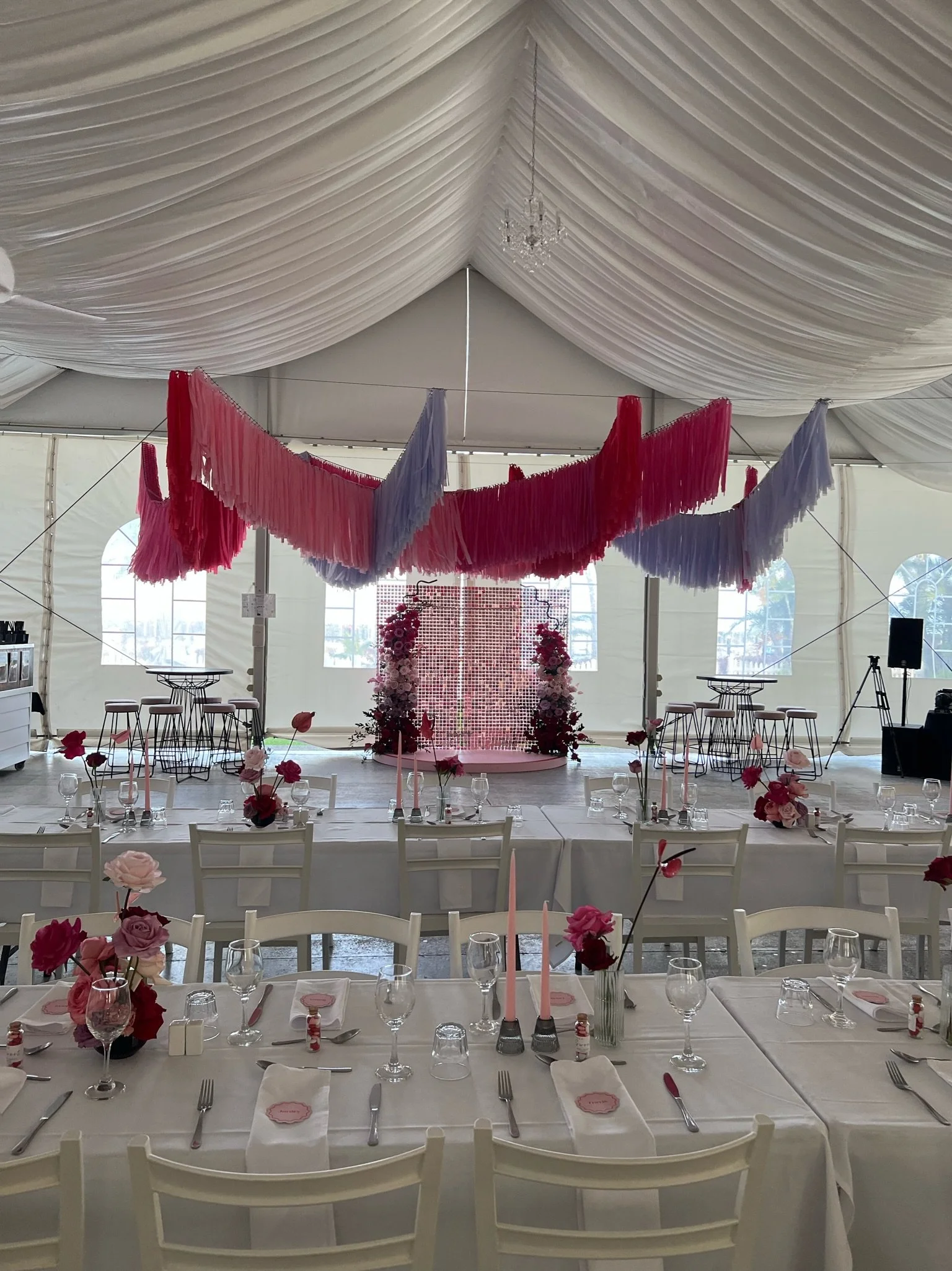 Elegant wedding reception setup inside a large tent with white draped ceiling, pink and purple hanging streamers, floral arrangements on tables, and a decorated backdrop at the front.