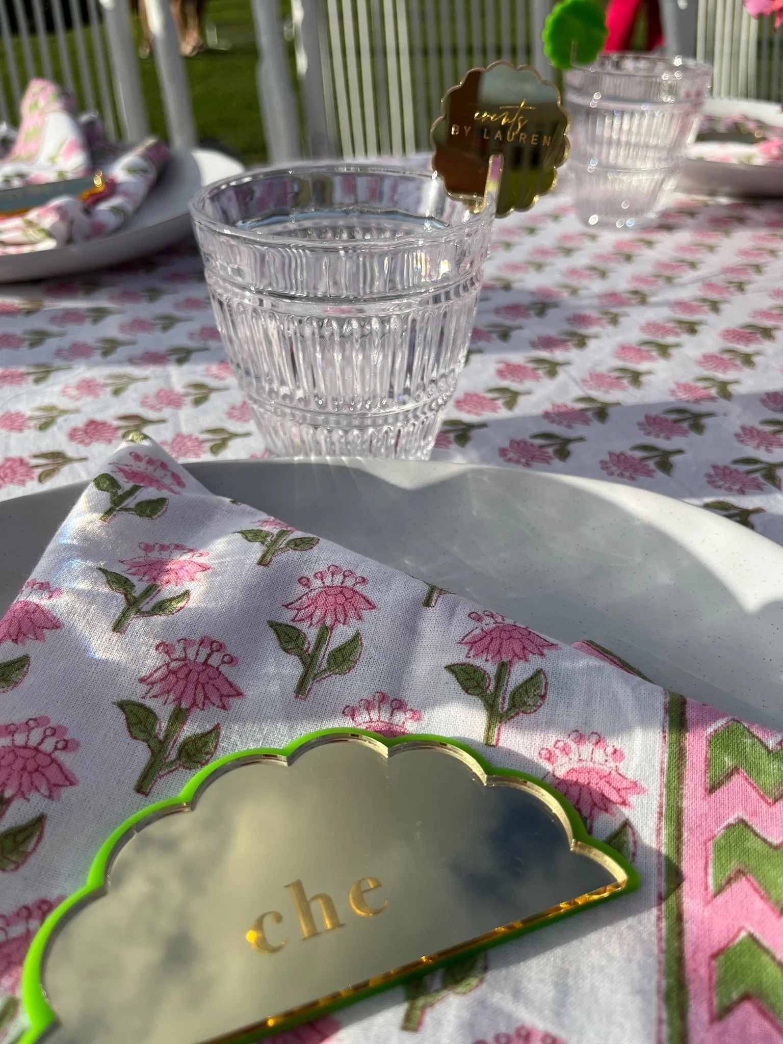 A clear glass of water with a 'Tiffany by Lauren' sticker on the rim, placed on a floral-patterned tablecloth at an outdoor gathering. In the foreground, there is a decorative glass and a cloth with pink flowers and green leaves. A small mirrored sig