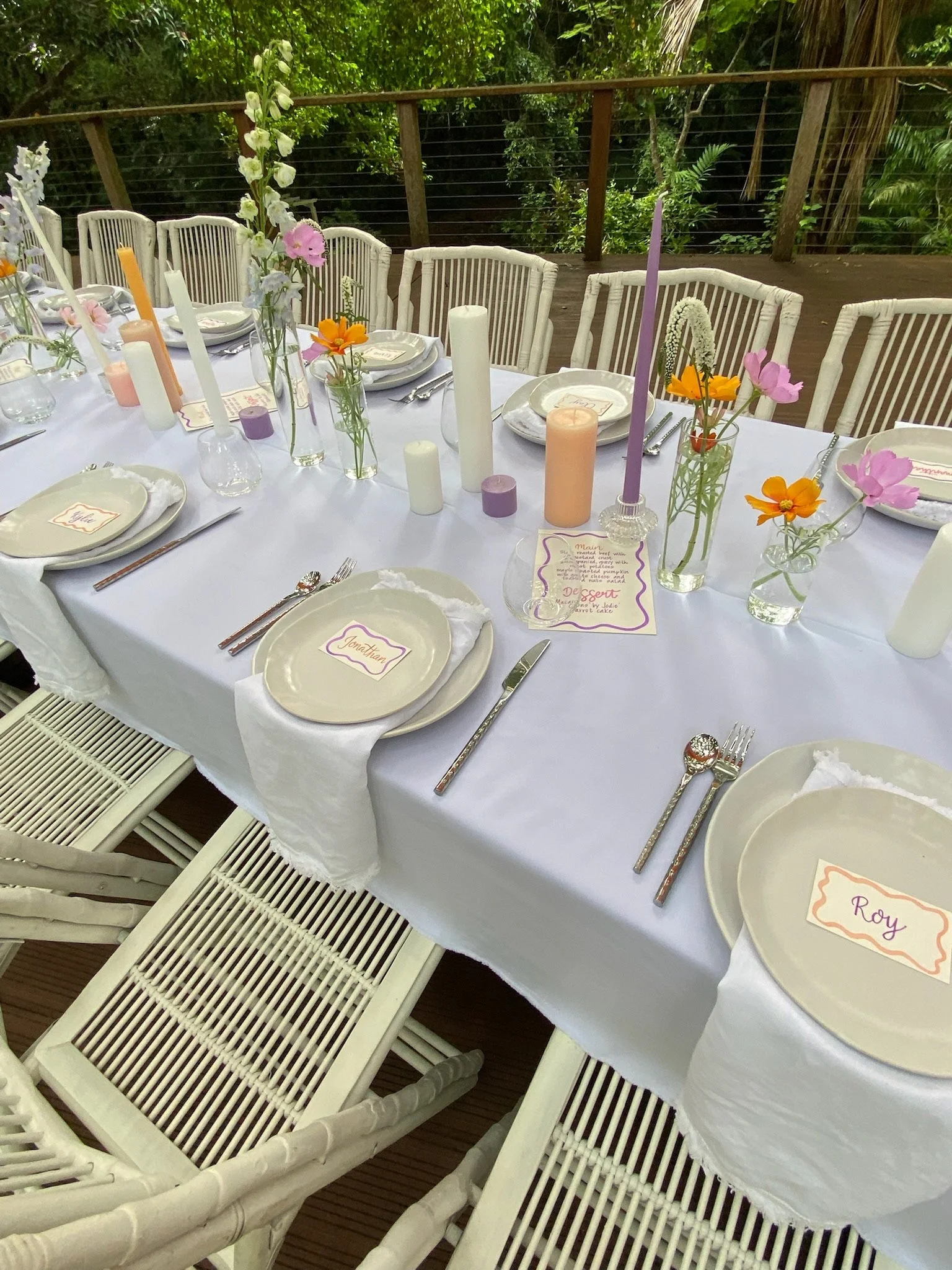 Decorative outdoor dining table set with white tablecloth, colorful flowers, candles, and place cards.