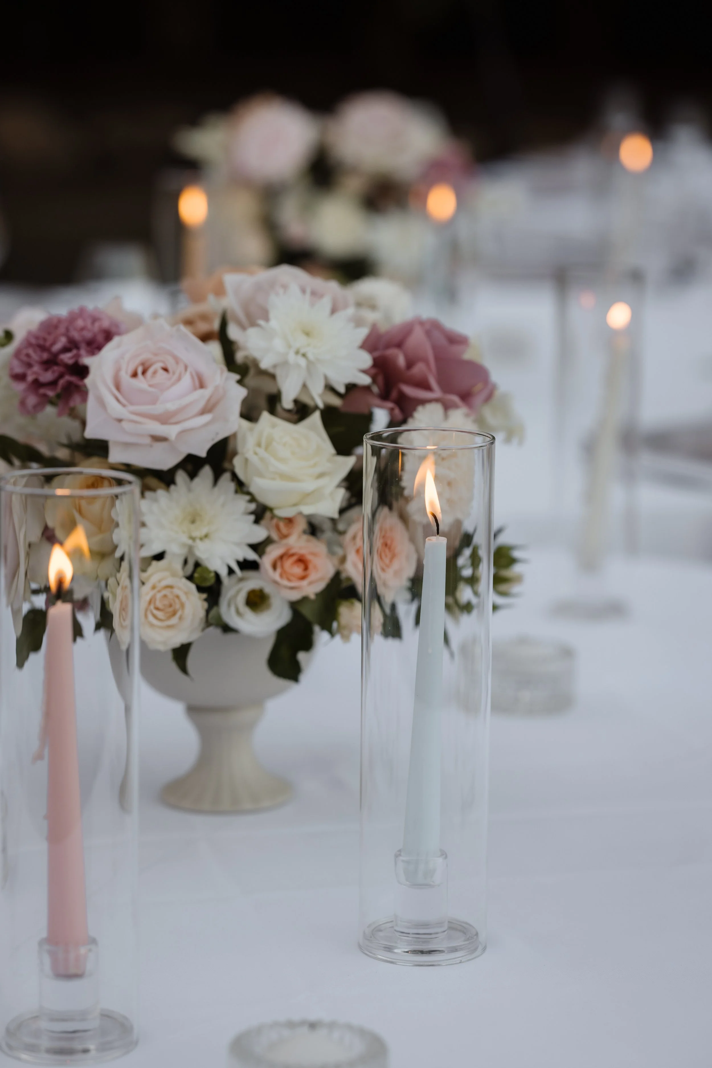 Wedding table decor with pink and white flowers in a white vase and candles in tall glass holders.