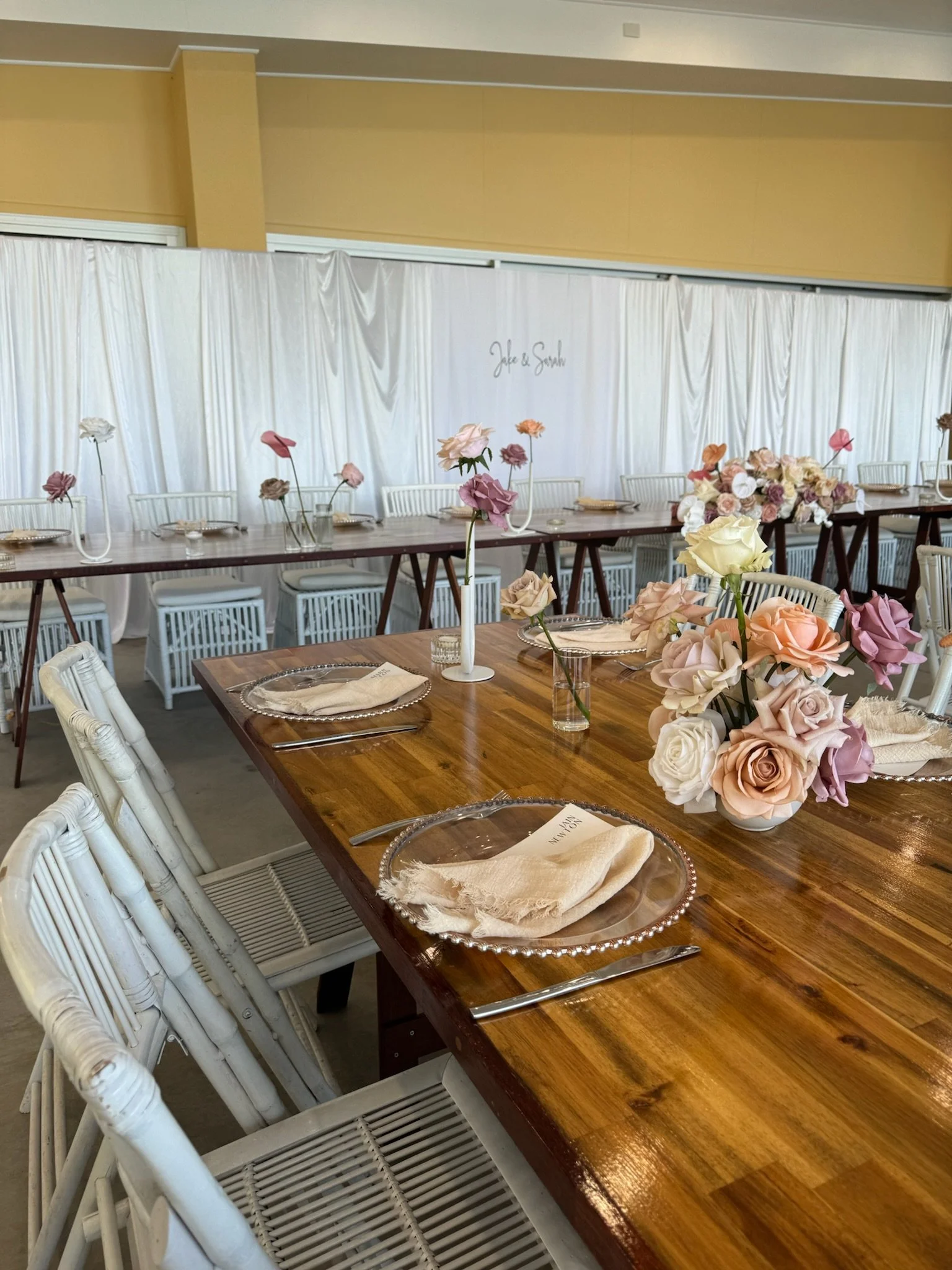 Decorated event space with long tables, floral centerpieces, and place settings, featuring soft pastel-colored roses and napkins.