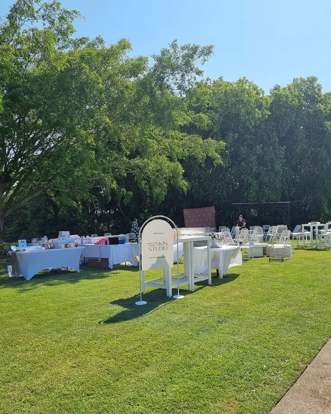 Outdoor event setup with tables display skincare products, surrounded by green trees on a sunny day.