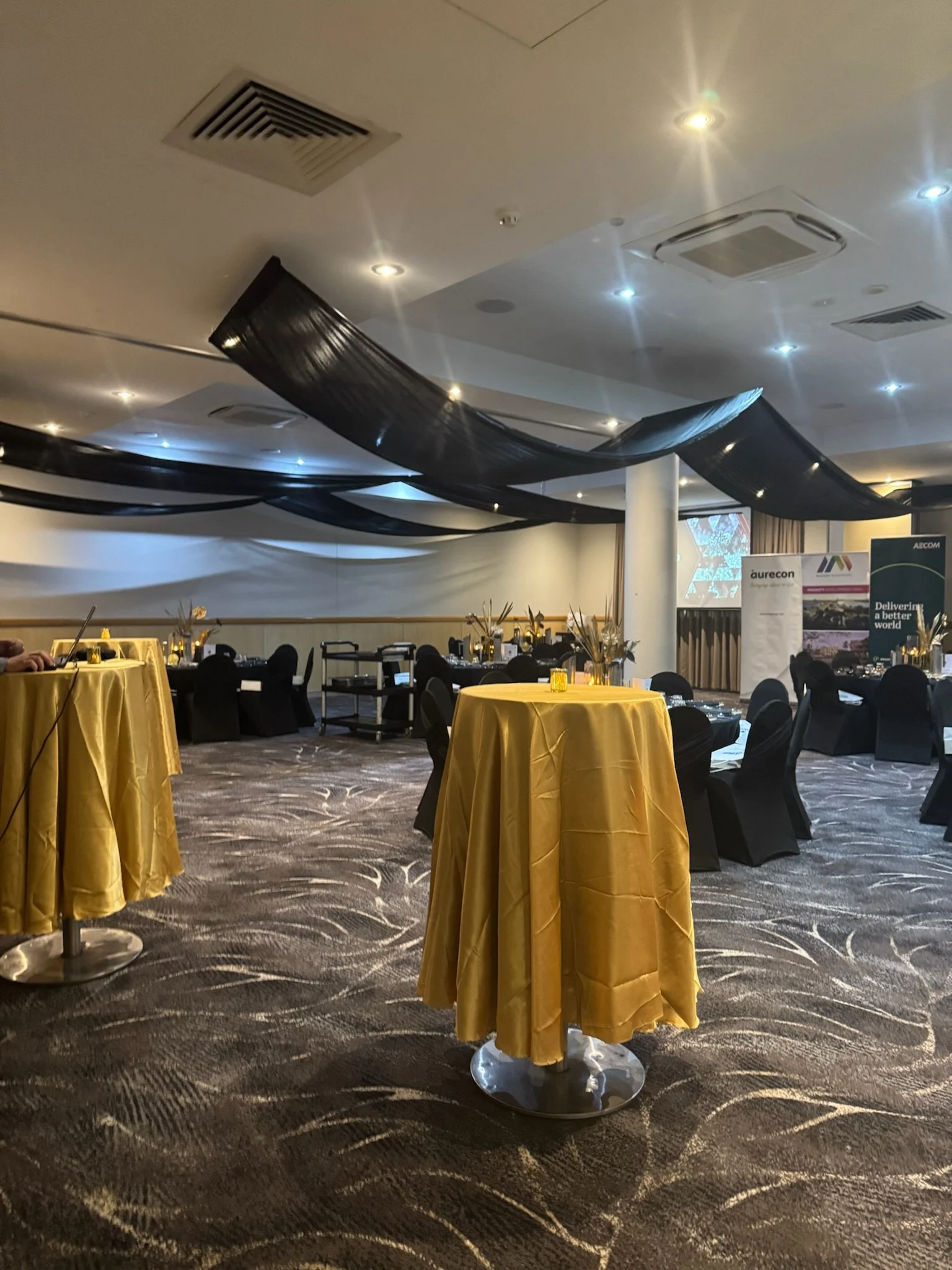 A decorated event hall with black and gold themed decor. There are round tables with yellow tablecloths and black chairs, and black fabric draped from the ceiling. The floor has a patterned carpet, and there are some screens and banners in the backgr