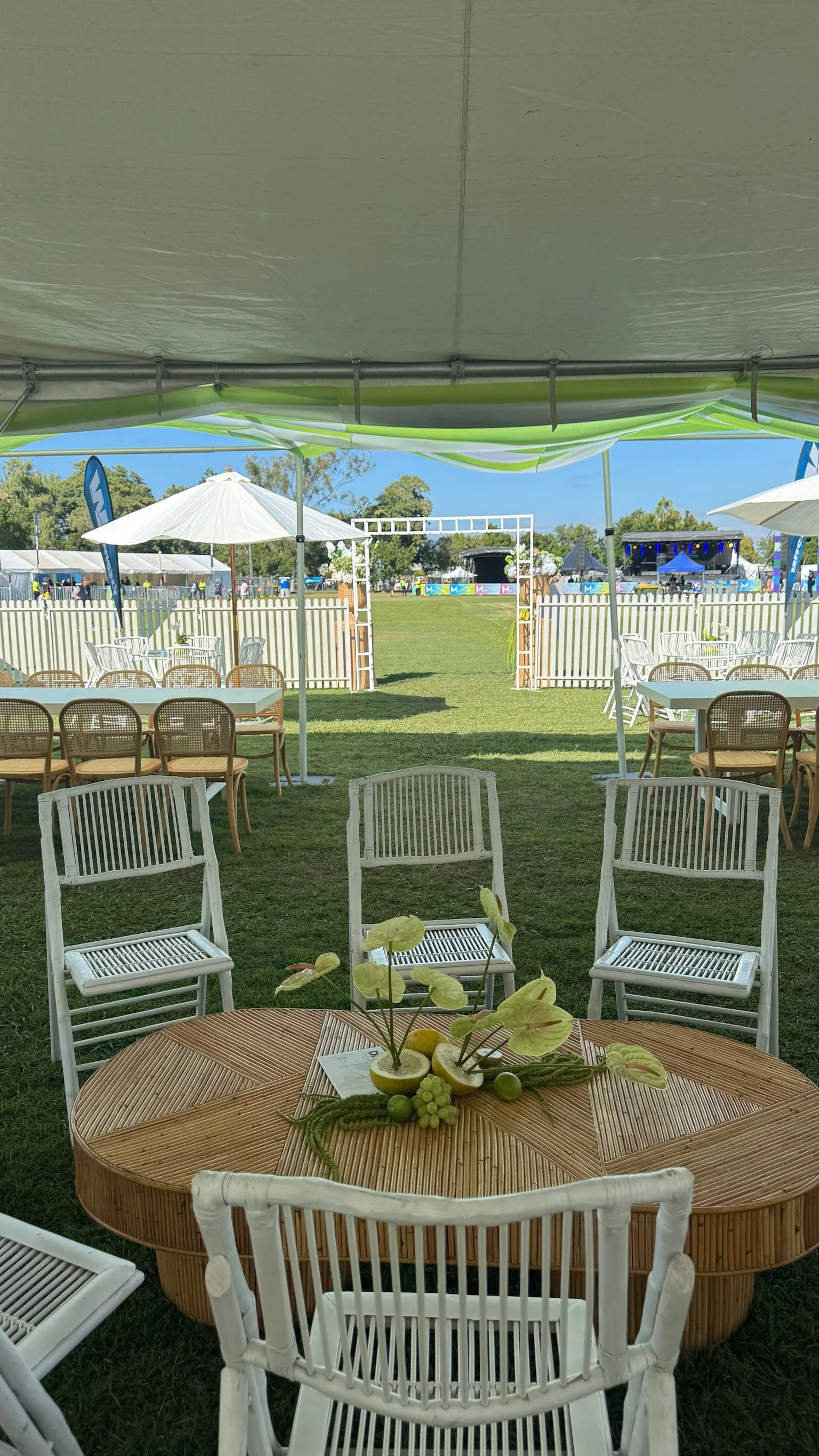 Outdoor event setup with white chairs, a round wooden table with a floral arrangement, tents, umbrellas, and a stage in the background.