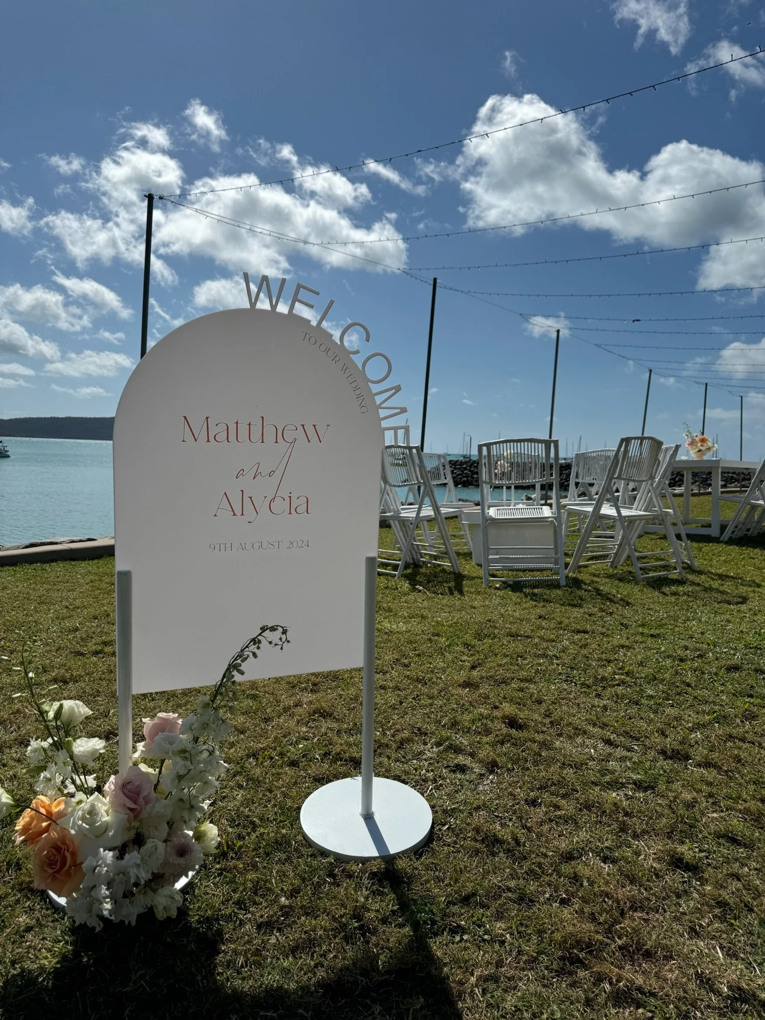Wedding sign with floral decoration in front of outdoor seaside wedding setup, including white chairs and string lights in the sky, on a grassy area near water and blue sky with clouds.
