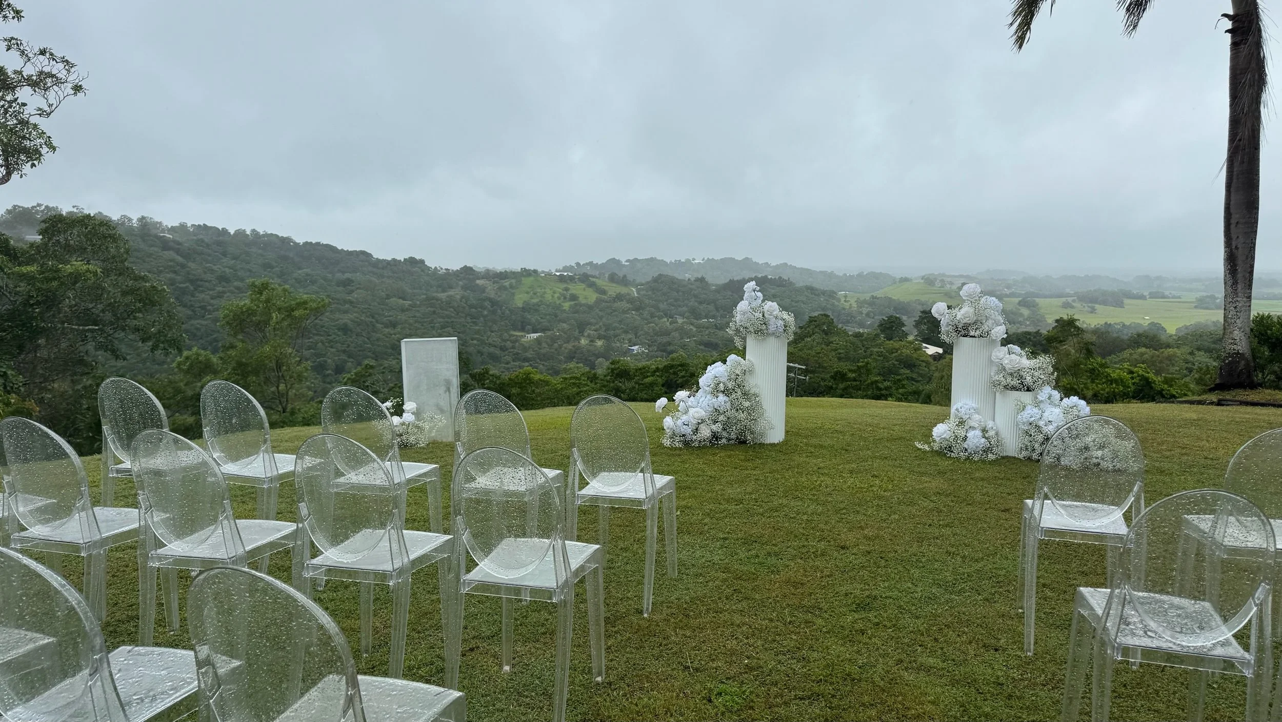 Outdoor wedding ceremony setup with transparent chairs and white floral arrangements on tall white pedestals, on a grassy field with rolling hills and trees in the background, under an overcast sky.
