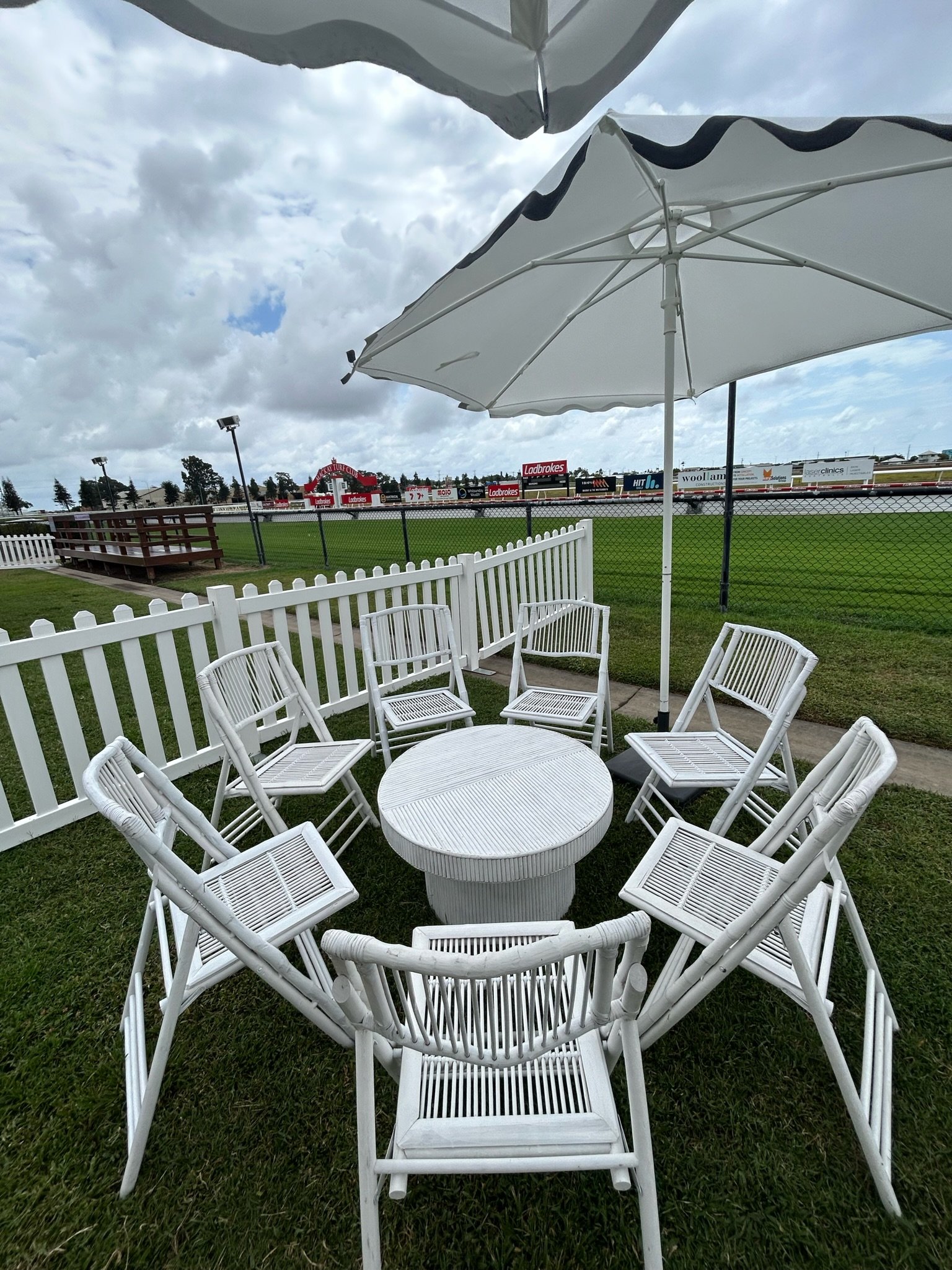 White outdoor patio furniture arranged in a circle with a small round table, under large white umbrellas, next to a white fence, overlooking a sports field with advertisements, on cloudy day.
