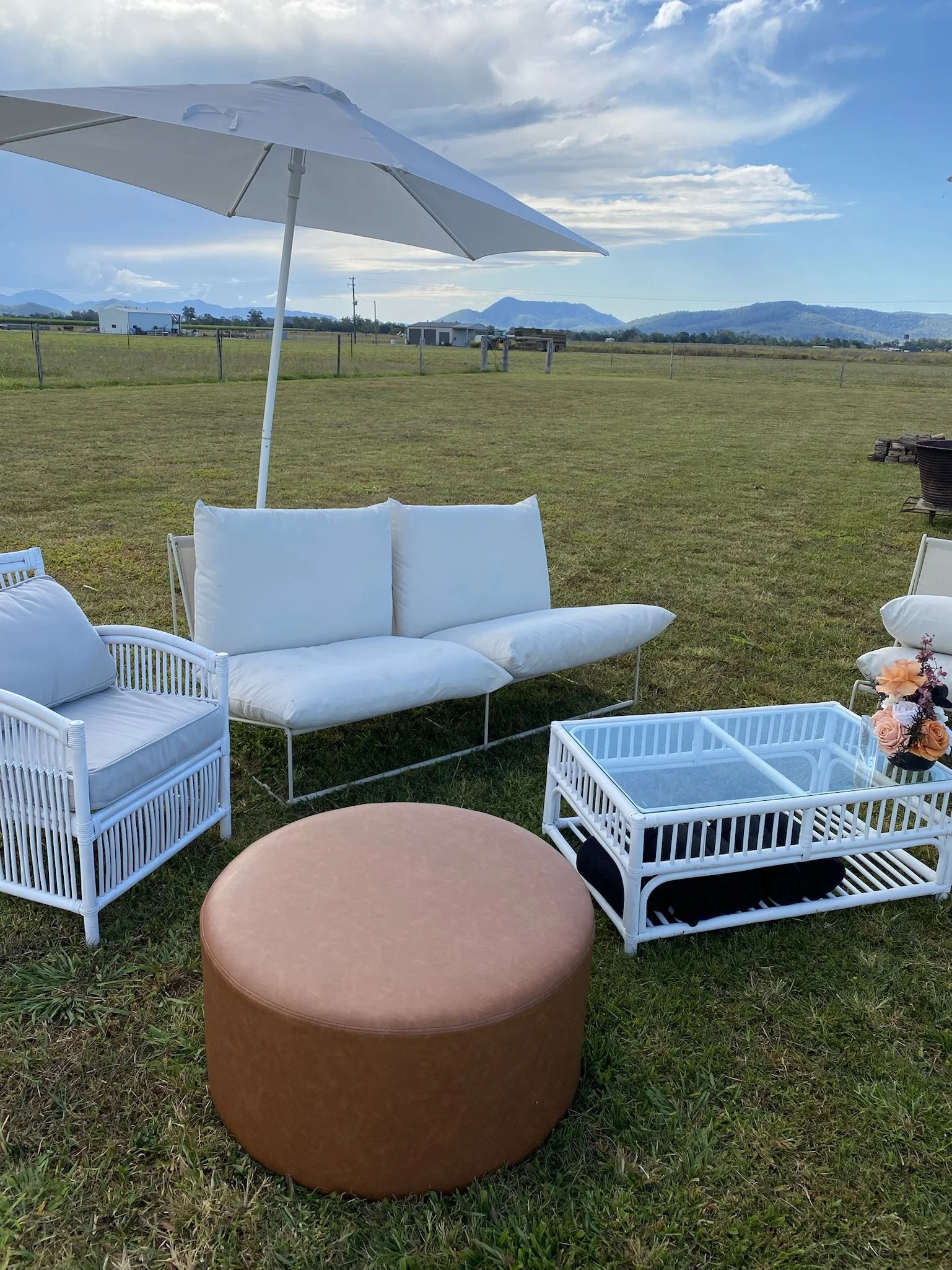 Outdoor seating area with white wicker chairs, a white cushioned sofa, a glass-top table, a large beige round ottoman, under a white umbrella, set in a grassy field with mountains and cloudy sky in the background.