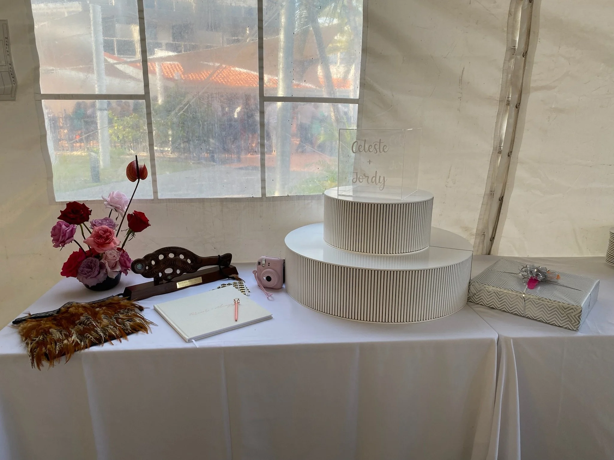 Decorated table with pink and red roses in a vase, a feathered accessory, a framed sign with names 'Celeste + Jordy', a pink instant camera, a spiral guestbook with a gold pen, a wrapped gift, and a small clear container with flowers, set in front of