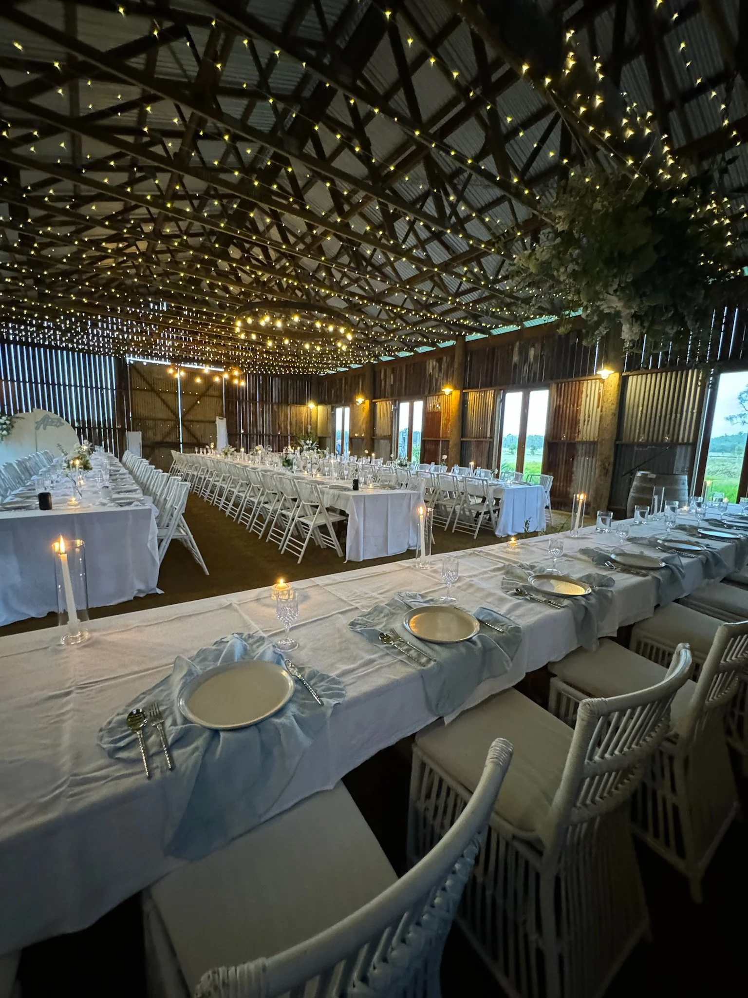 Wedding reception setup inside a barn with long dining tables covered in white tablecloths, decorated with candles and tableware, illuminated by string lights and hanging greenery.