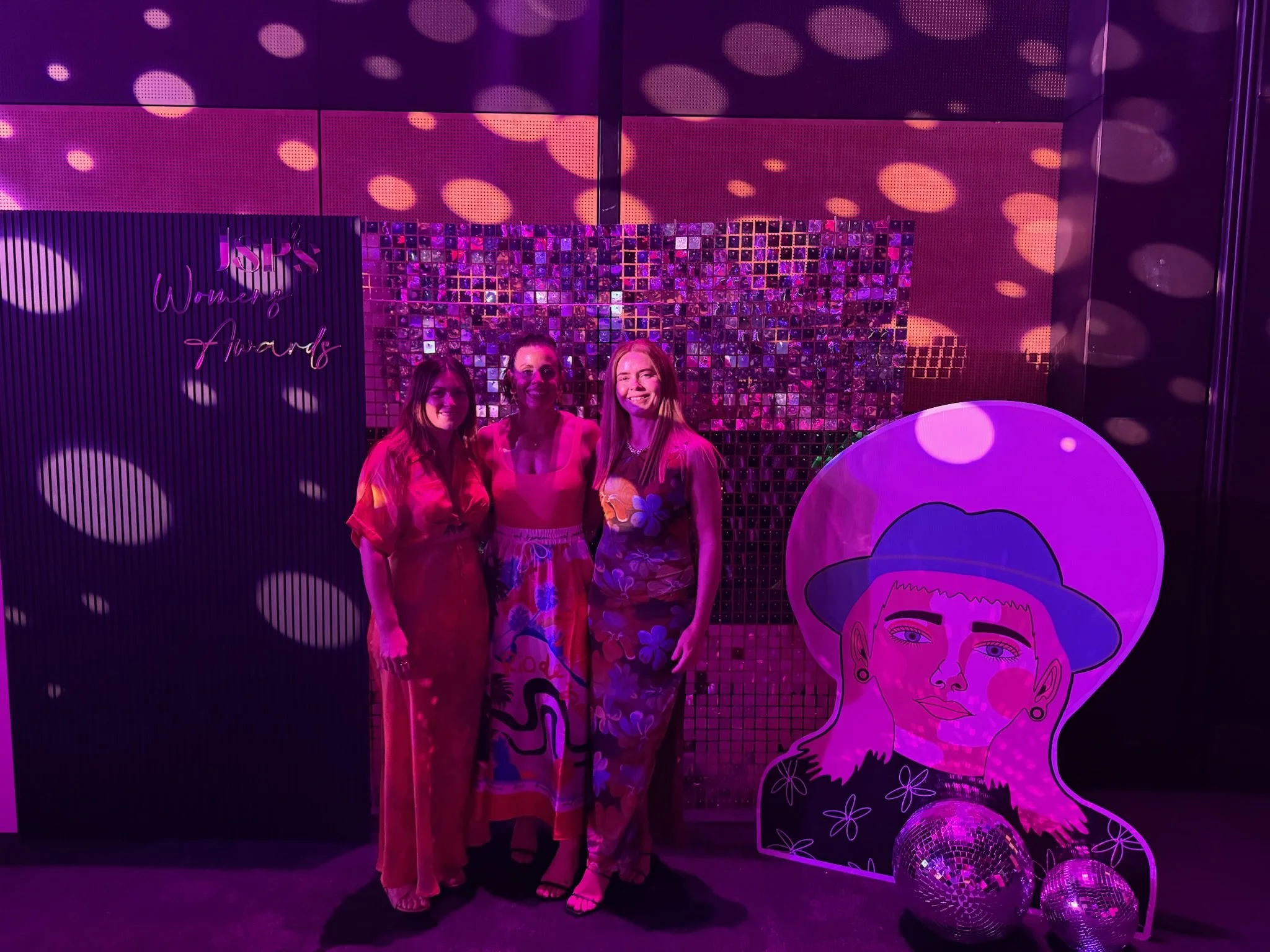 Three women in colorful clothing pose and smile at a women's awards event with a glamorous backdrop, artistic display, and disco balls.