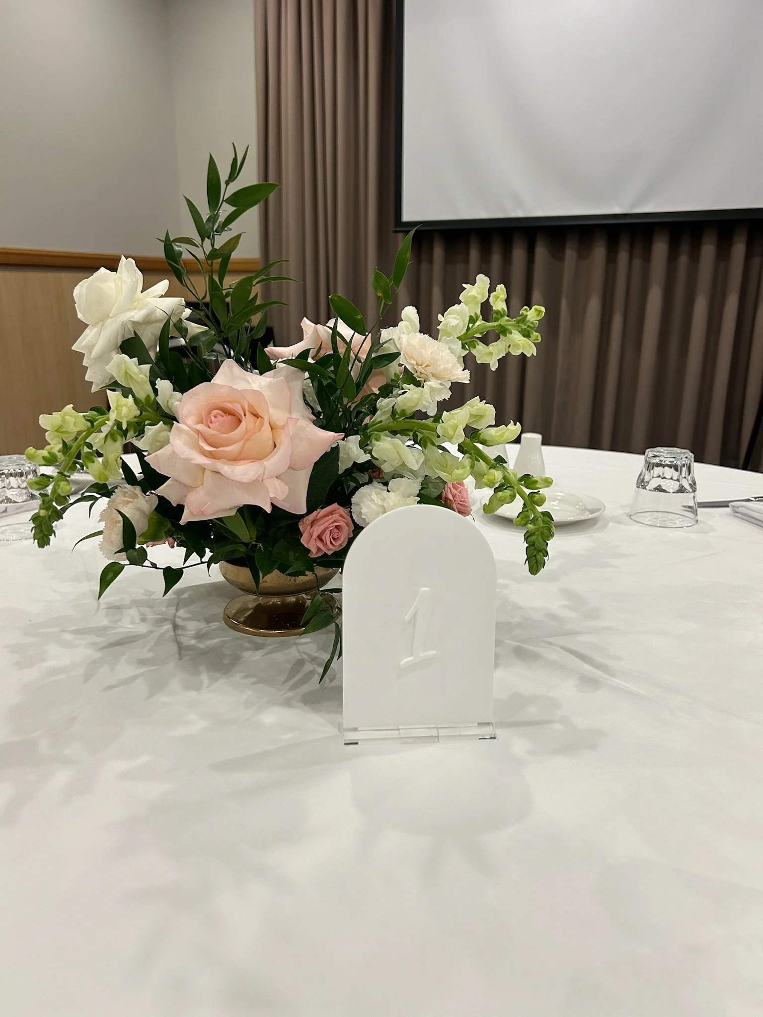 Elegant table centerpiece with pink and white flowers, set on a white tablecloth at an event or banquet, with a table number card labeled '1' in front.