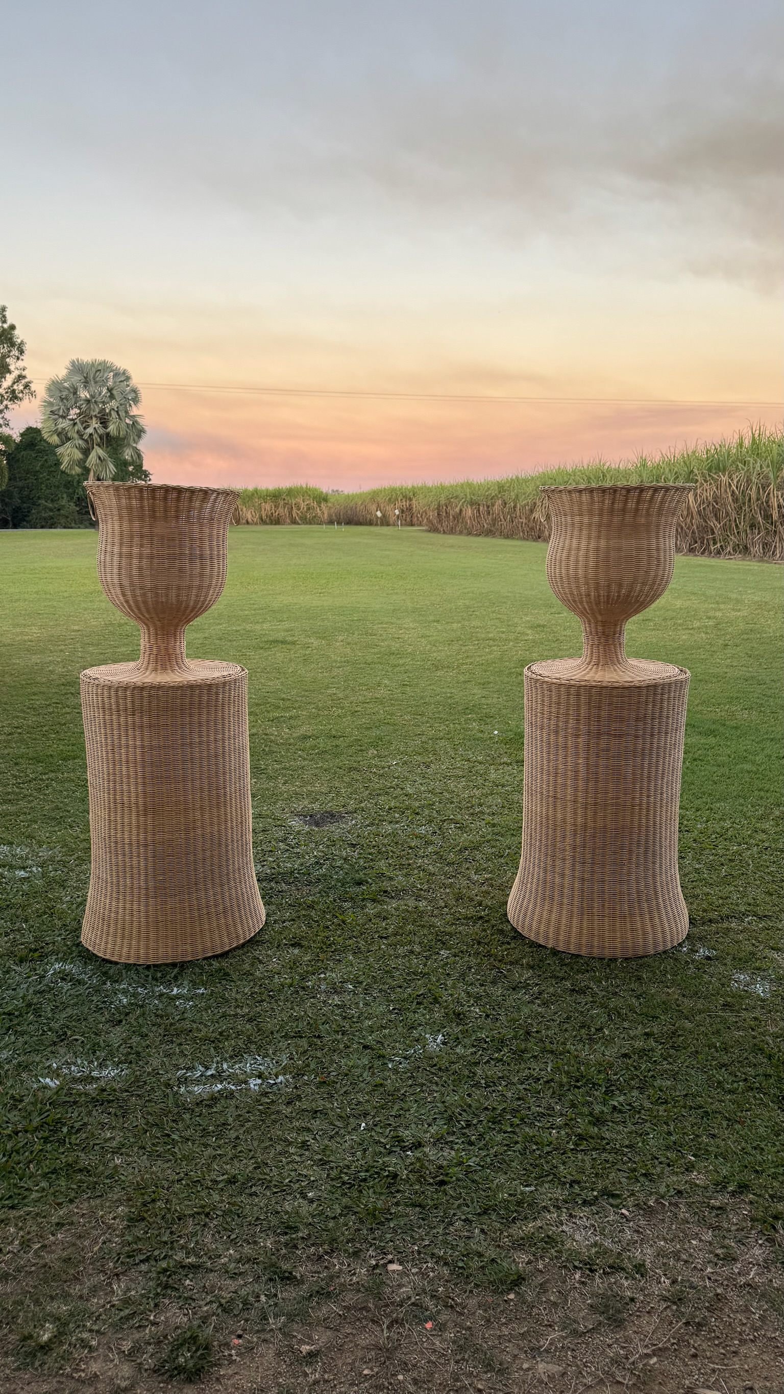 Two tall wicker planters on the grass in an open field at sunset with a pink and orange sky and trees in the background.