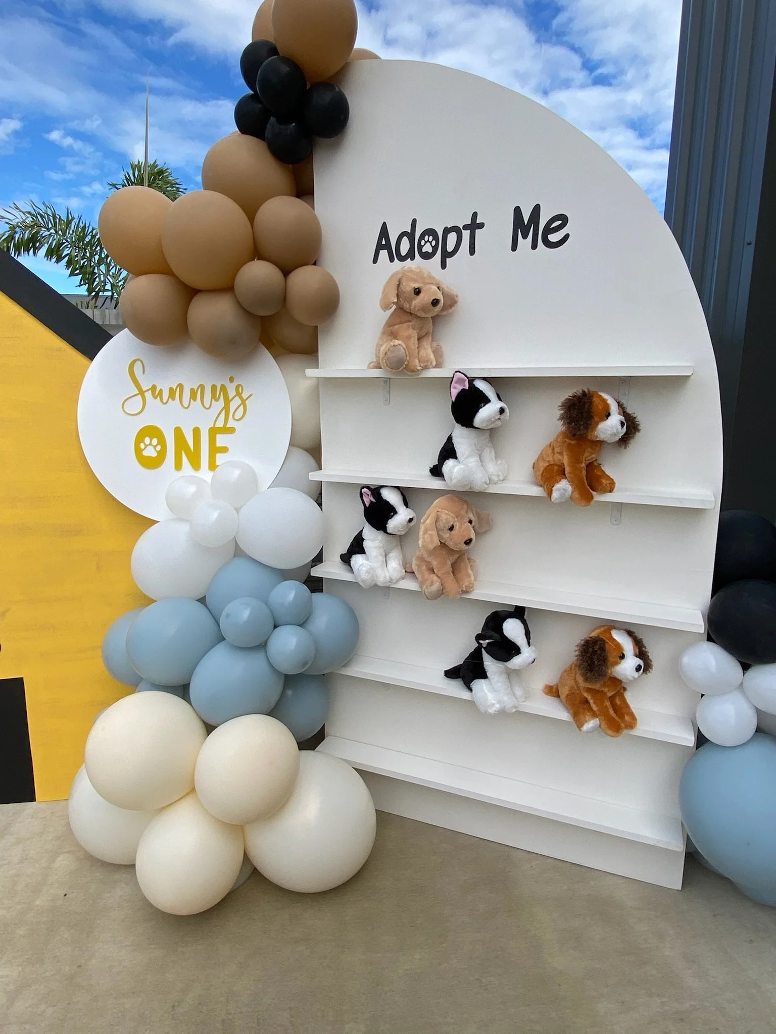 A pet adoption display with plush dogs and cats on white shelves, surrounded by balloon decorations in brown, black, white, blue, and cream, with signs reading 'Adopt Me' and 'Sunny's ONE'.
