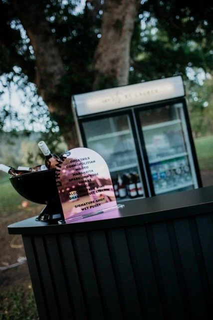 A black outdoor bar table with a black bowl holding white glow sticks, a menu card, and a small wine bottle. In the background, there is a glass-front refrigerator with drinks, set outdoors under a tree.