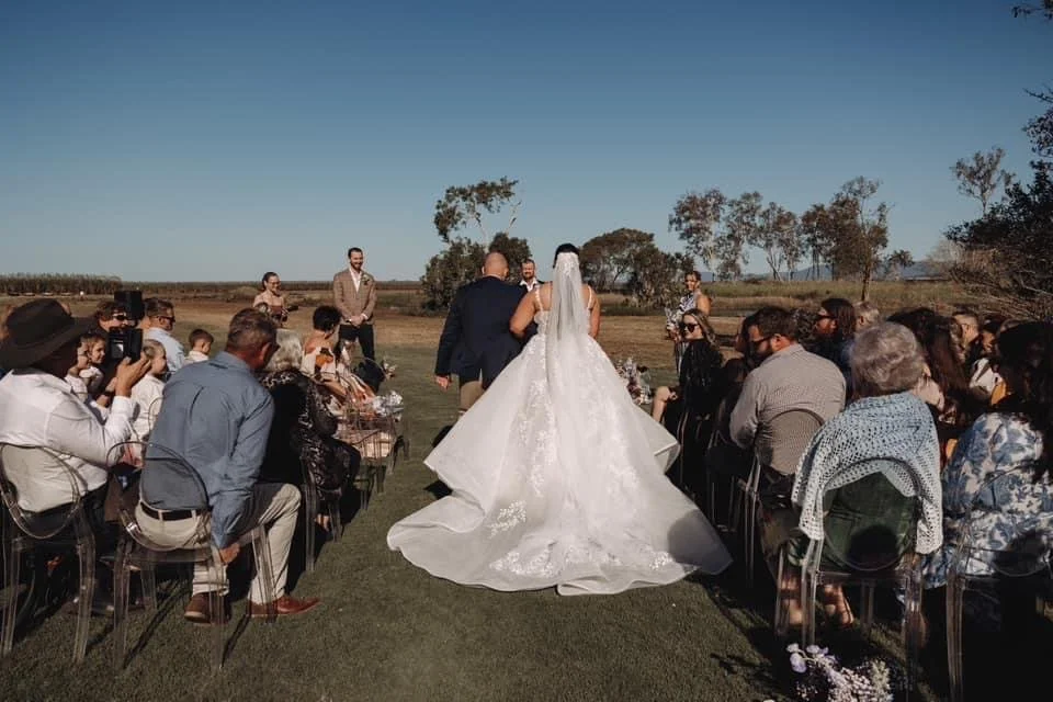 Wedding ceremony outdoors with a bride in a white gown and veil walking down the aisle with the groom, surrounded by seated guests on a grassy field under a clear sky.