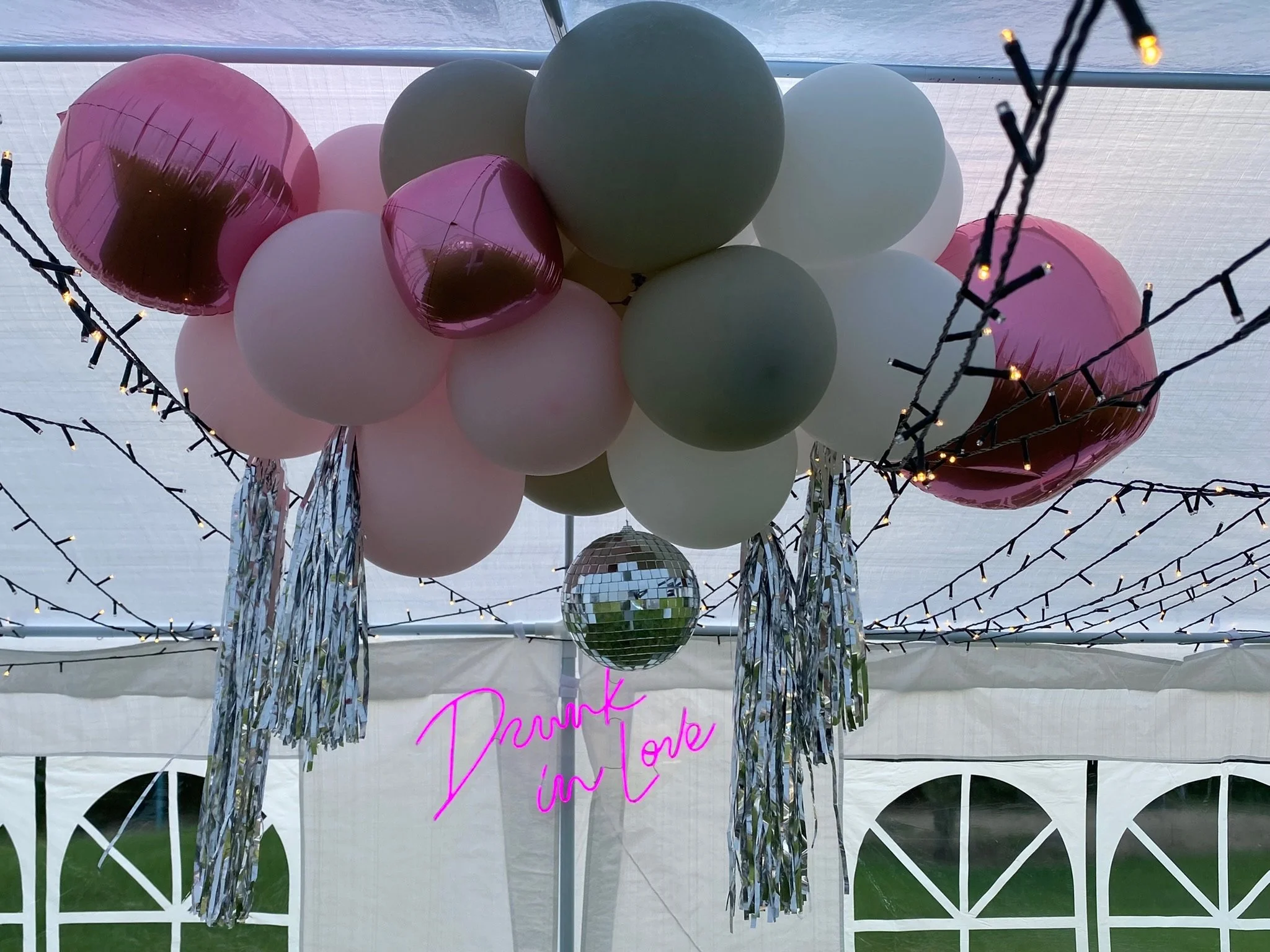 Decorative balloons in pastel colors and metallic balloons hanging from a string of small fairy lights, with a neon sign that says 'Drunk in Love' attached to the balloon arrangement, in a tent.