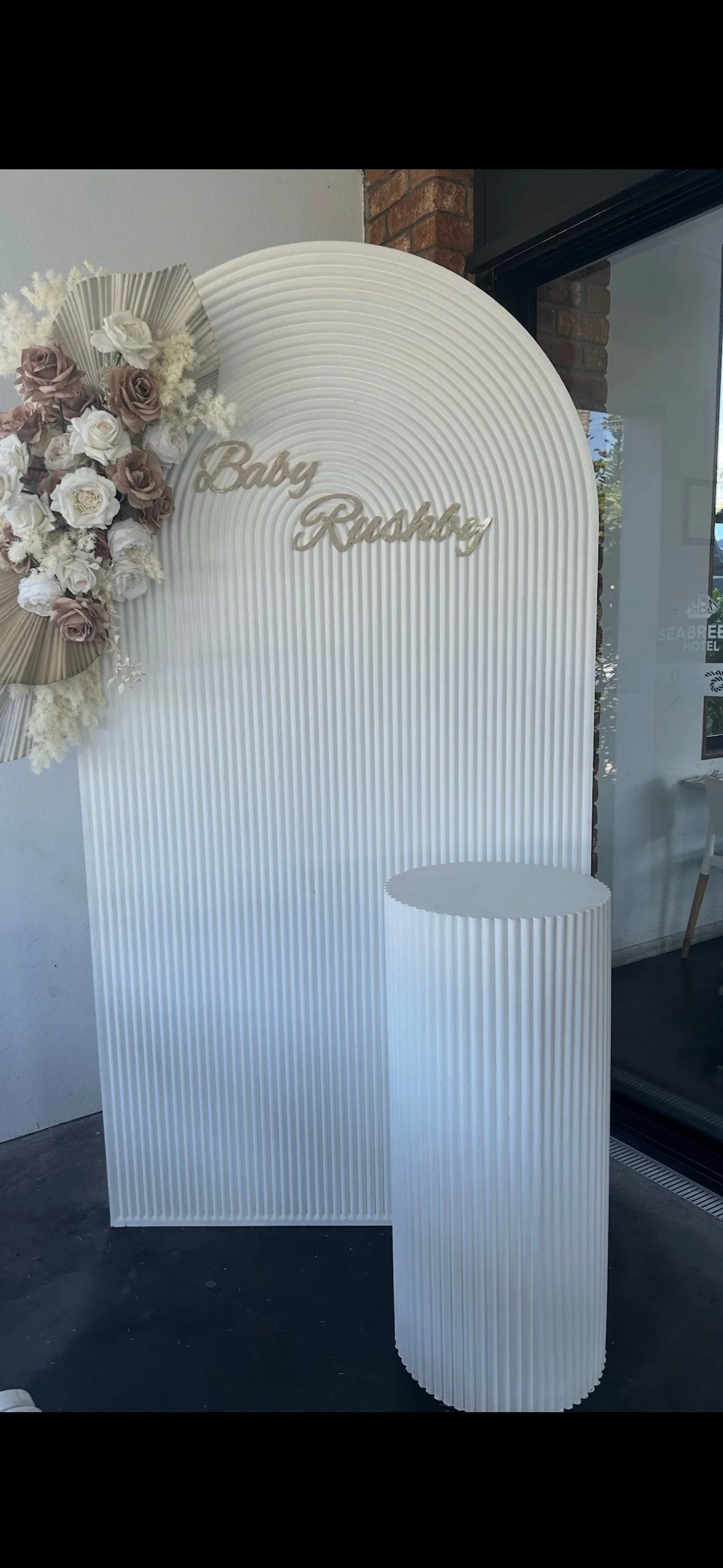 Decorative white backdrop with vertical ridges and a curved top, decorated with a floral arrangement of white and beige roses and pampas grass, with the words "Baby Rushed" in metallic cursive. A matching white, ridged cylindrical stand is placed in 