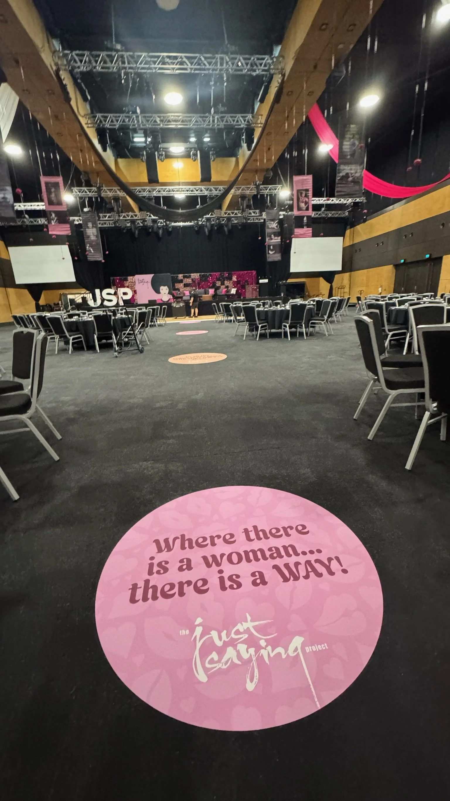 Empty event hall with round tables and chairs, stage with pink and black decorations, and a large pink circular marker on the floor reading 'Where there is a woman... there is a WAR! the 'Just Saying' project.'