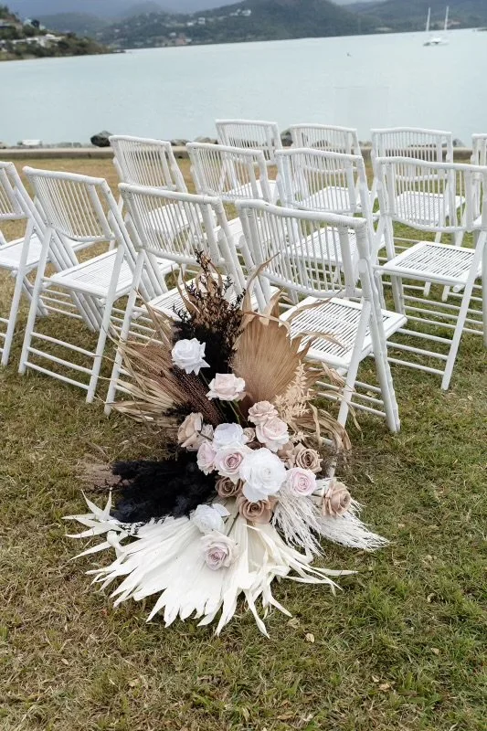 Empty white chairs arranged outdoors near a body of water, with a large floral arrangement featuring white and blush roses, dried leaves, and black accents in the foreground.