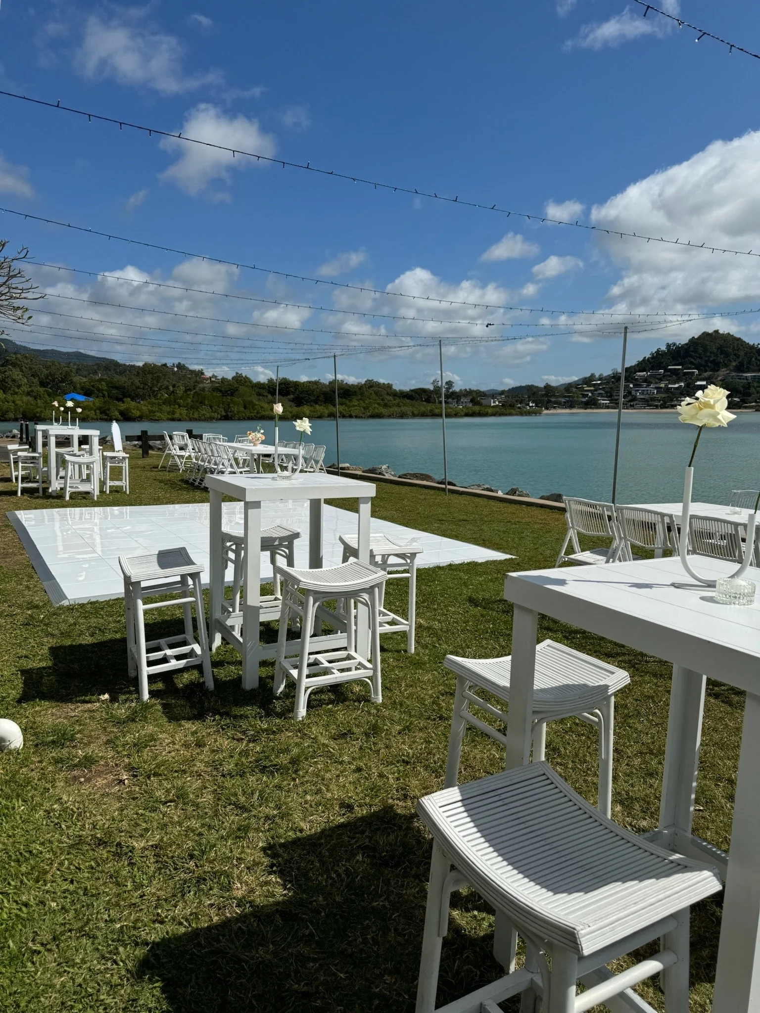 Outdoor event setup near water with white tables and chairs, decorated with flowers, under blue sky with clouds, string lights overhead.