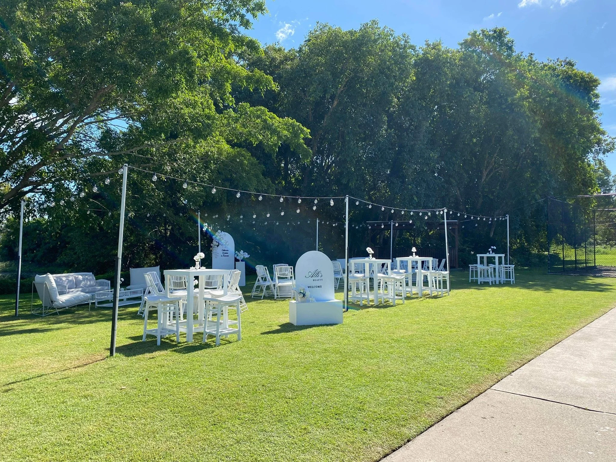 Outdoor event setup with white furniture, tables, and string lights on a grassy area with large green trees in the background.