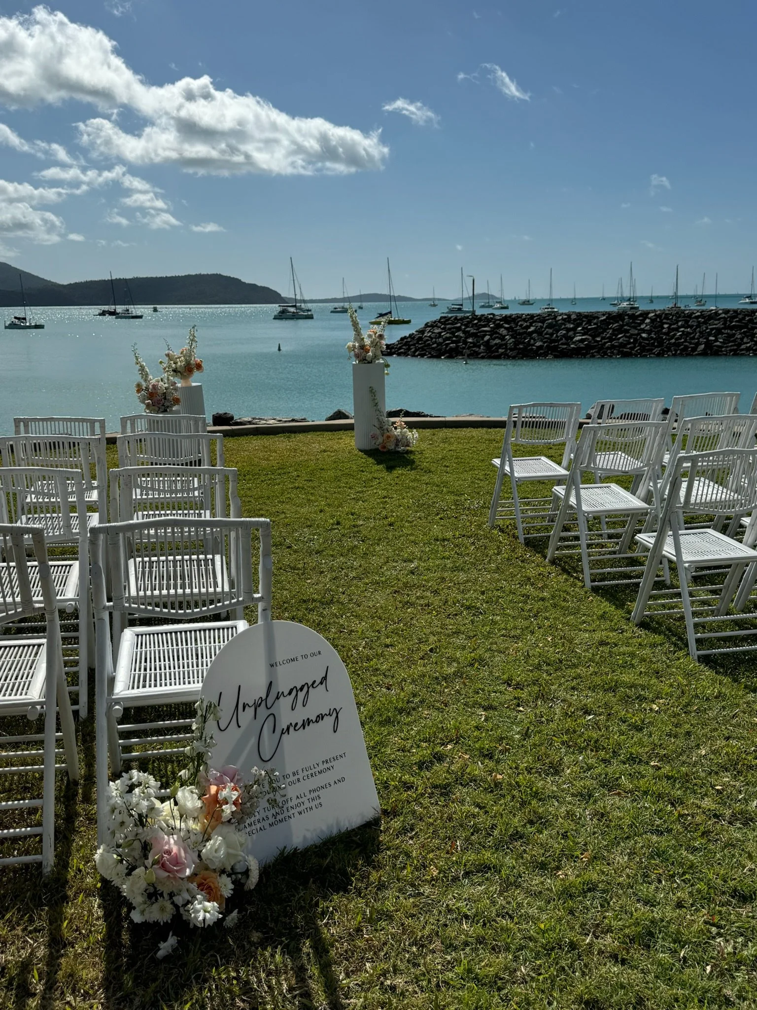 Seaside wedding setup with white chairs arranged on green grass, floral decorations, and a sign that reads 'Unplugged Ceremony' near the water, with sailboats and a breakwater in the background under a partly cloudy sky.