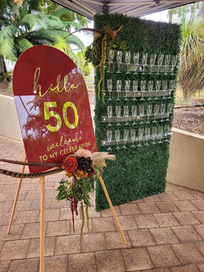 A decorated sign with a red background and gold lettering saying "hello 50" and "welcome to my celebration" next to a green artificial hedge wall with multiple empty champagne glasses displayed on it, under a canopy outdoors with tropical plants in t