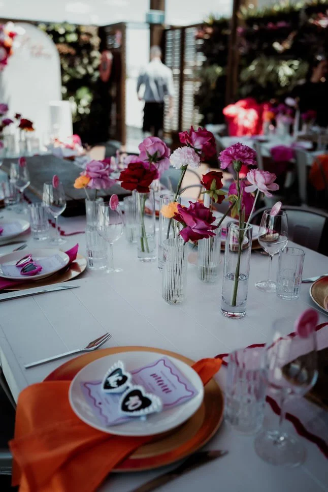 A beautifully decorated outdoor dining table with pink, red, and white flowers in glass vases, set with plates, wine glasses, and utensils, with a person in a white shirt and black pants in the background.