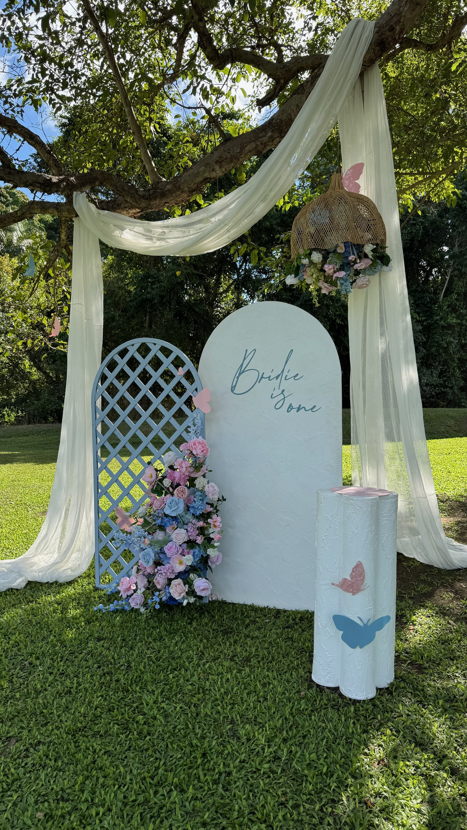 Outdoor wedding ceremony setup with a white arch draped in sheer fabric, decorated with pink, purple, and blue flowers, a white standing backdrop with 'Bride x One' written, a lattice panel, a floral arrangement, and butterfly cutouts on the backdrop