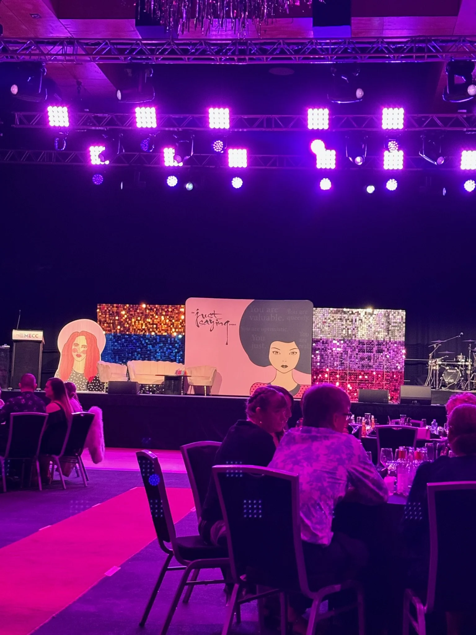 Stage with colorful artwork, purple lighting, and a seated audience at tables in a banquet hall.