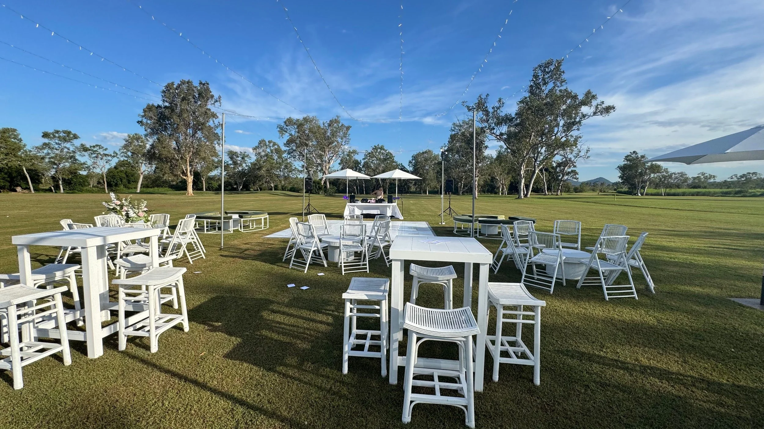 Outdoor event setup with white tables, chairs, and umbrellas on a grassy field, with string lights overhead and trees in the background.