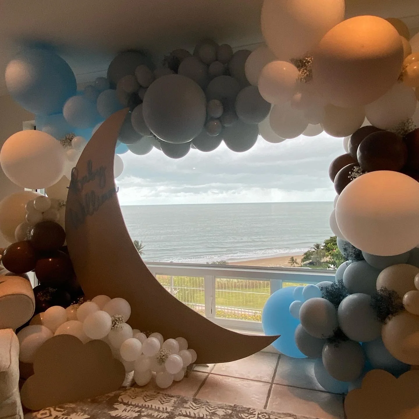 Decorative balloon arrangement with a large crescent moon shape, set against a window view of the ocean and beach.