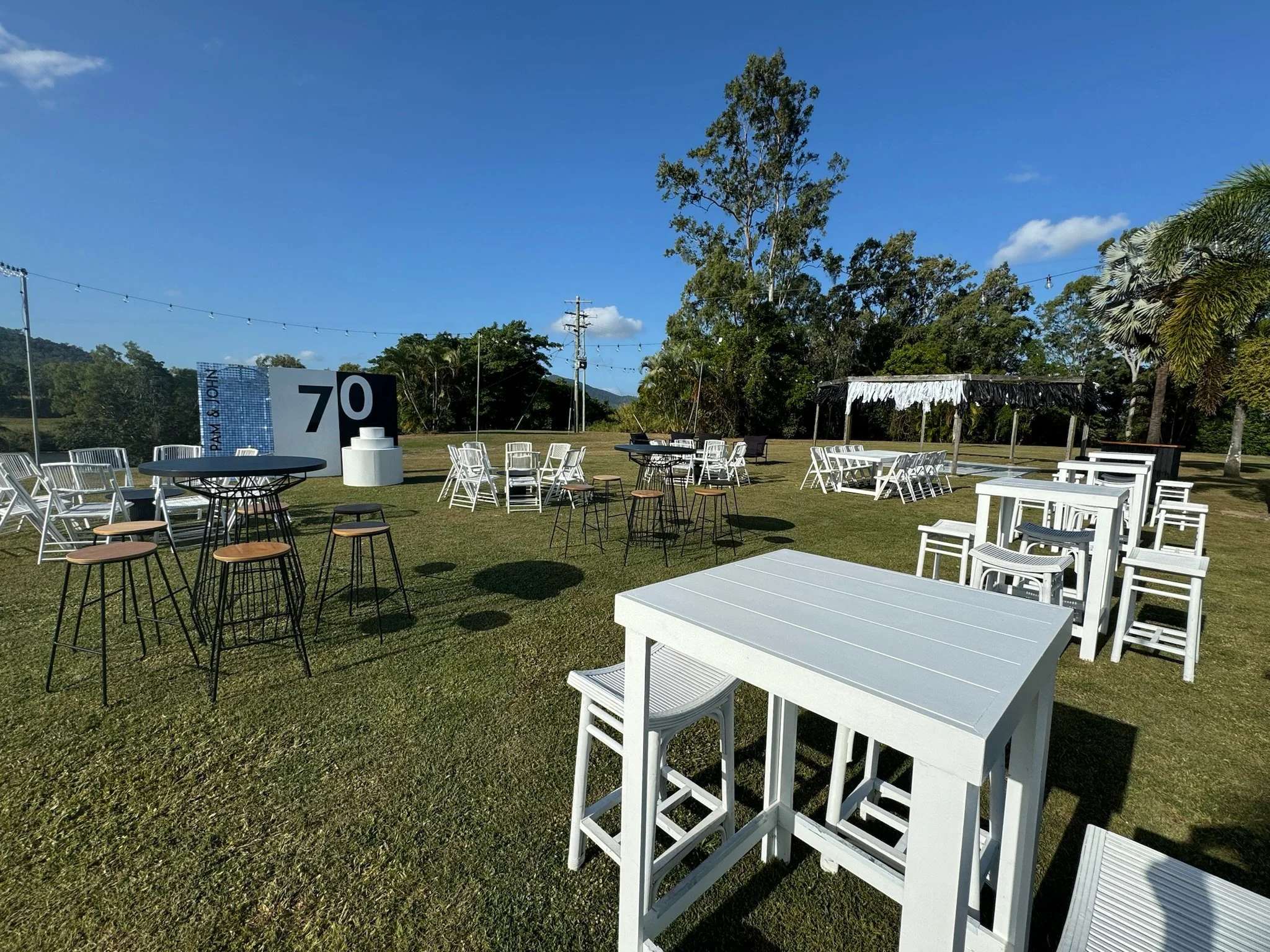 Outdoor event setup with various white and black tables and chairs on a grassy field under a blue sky with some clouds, surrounded by trees and a few string lights.