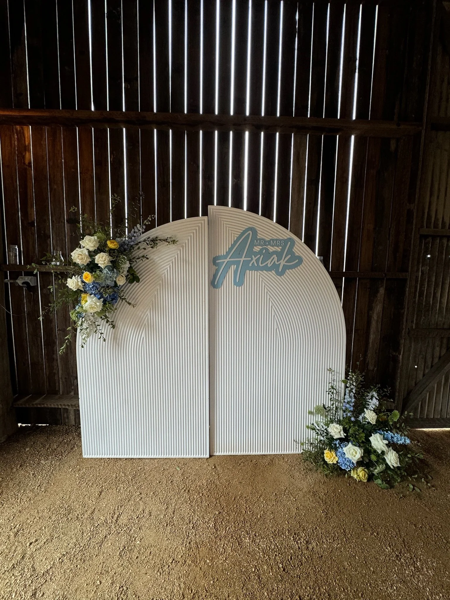 Wedding backdrop with floral arrangements and sign reading 'Mr. & Mrs. Axial' in a rustic barn setting.